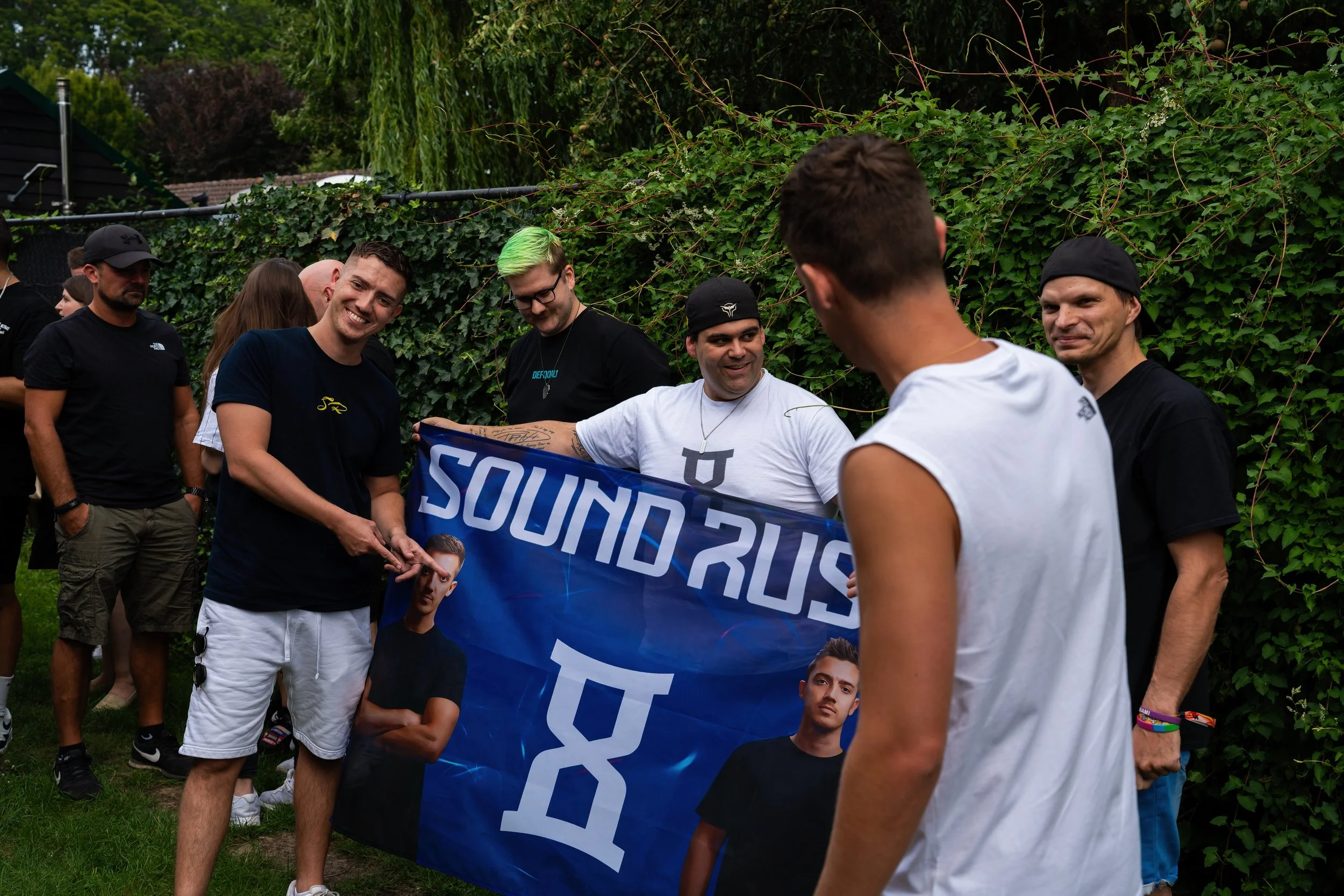 Gruppe von jungen Männern beim Treffen im Freien, einige halten ein Banner mit dem Schriftzug „SOUND RUSH“ und Bildern von DJs.