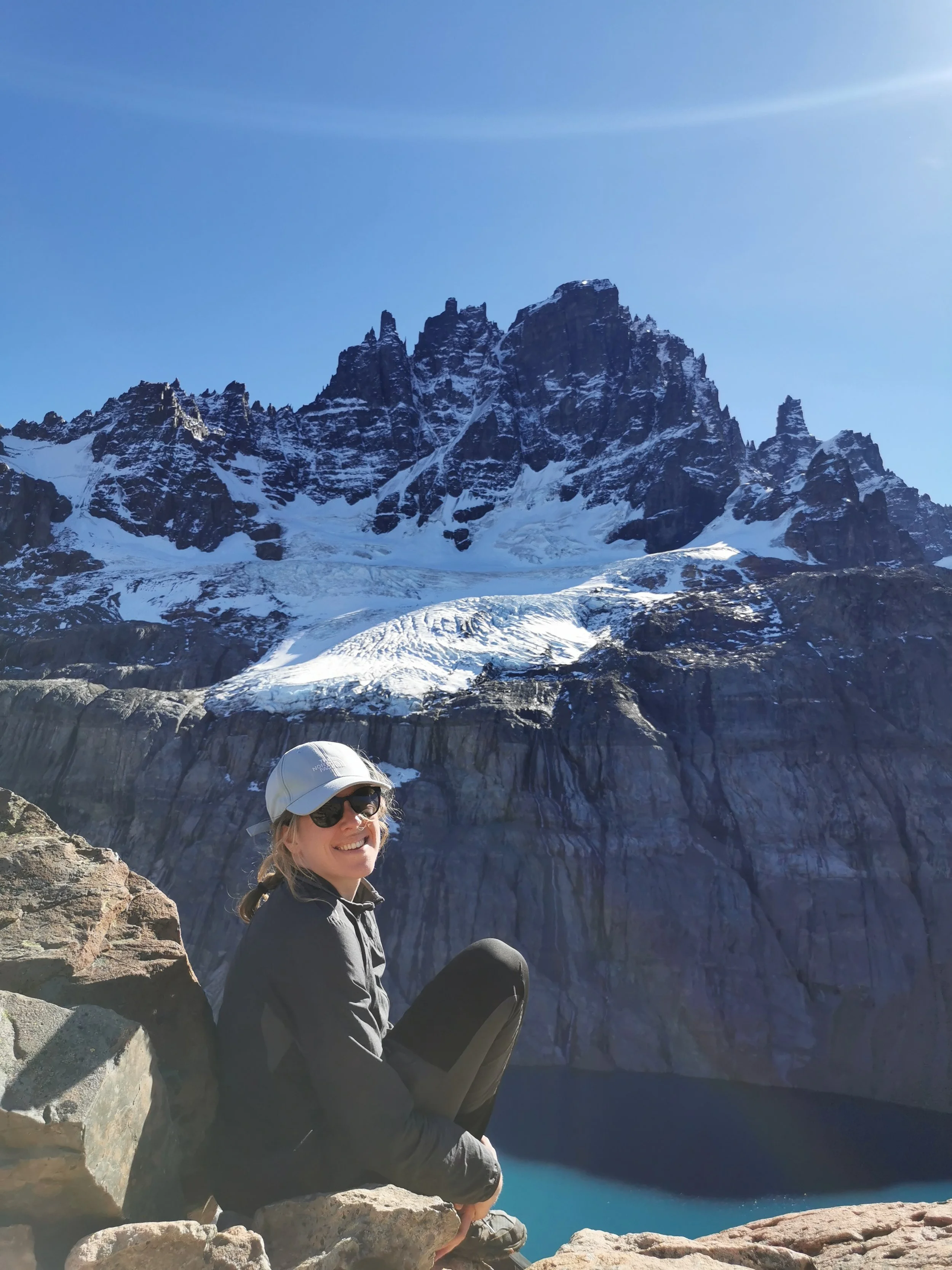 Une femme souriante portant des lunettes de soleil, un chapeau blanc et une veste noire, assise sur des rochers en face d'un lac bleu entouré de montagnes enneigées sous un ciel ensoleillé.