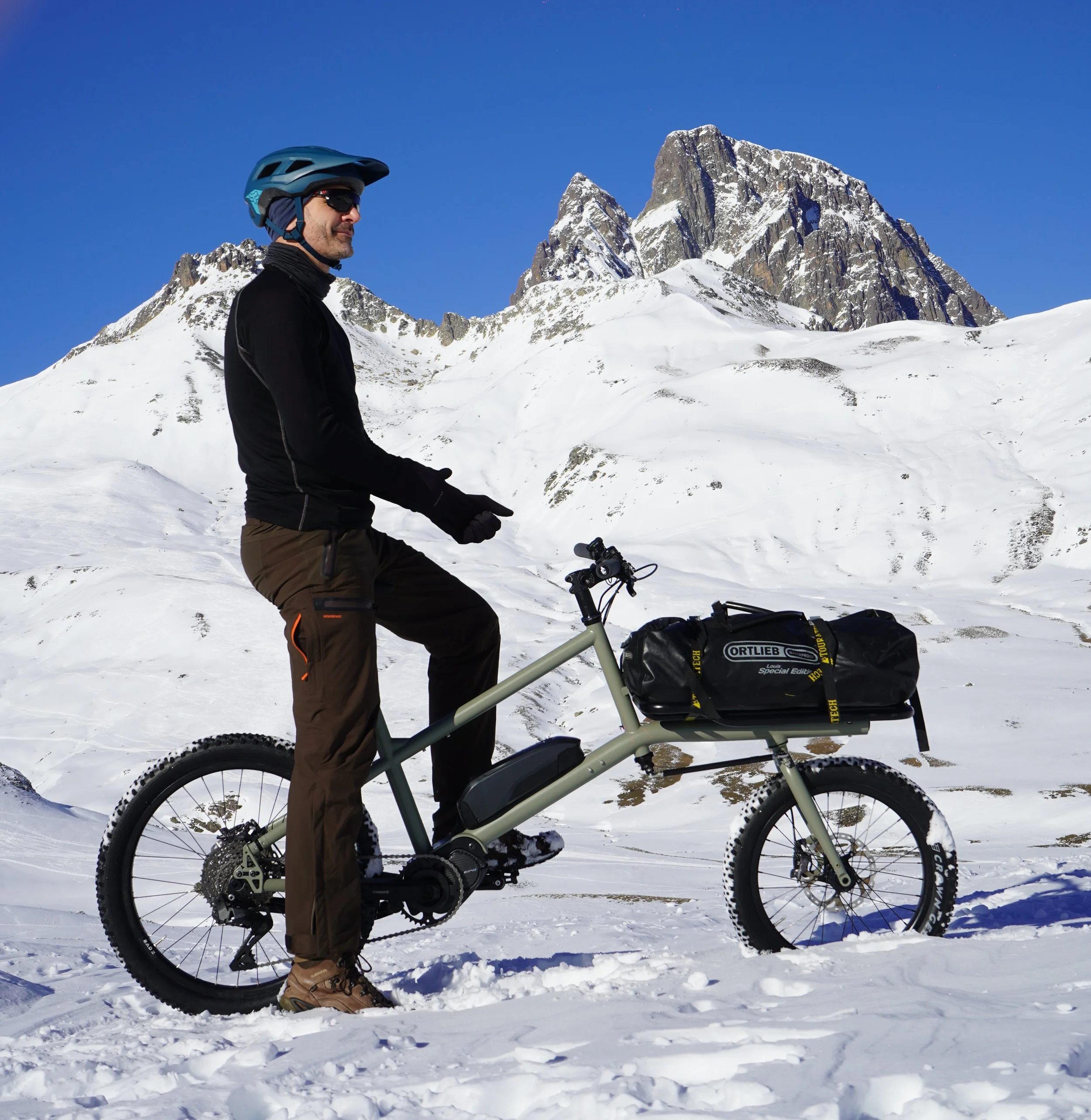 Homme en équipement de ski électrique à côté d'un vélo électrique dans un paysage enneigé avec montagnes en arrière-plan.