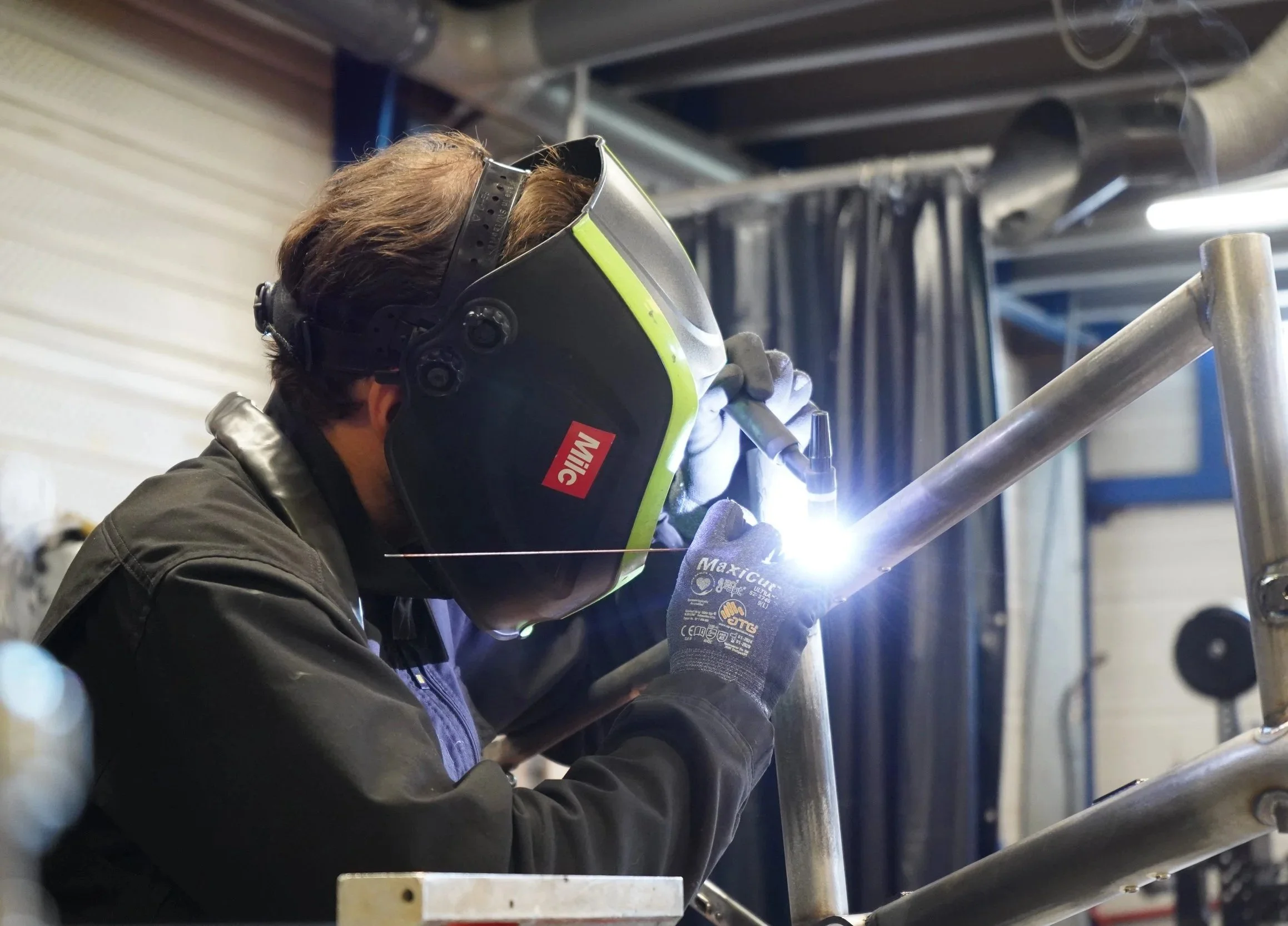 Un ingénieur effectue un soudage sur une structuren métalliques dans un atelier de fabrication, portant un casque de soudage avec le logo Maxic, des gants de protection, et utilisant un poste de soudage pour assembler un tube métallique.