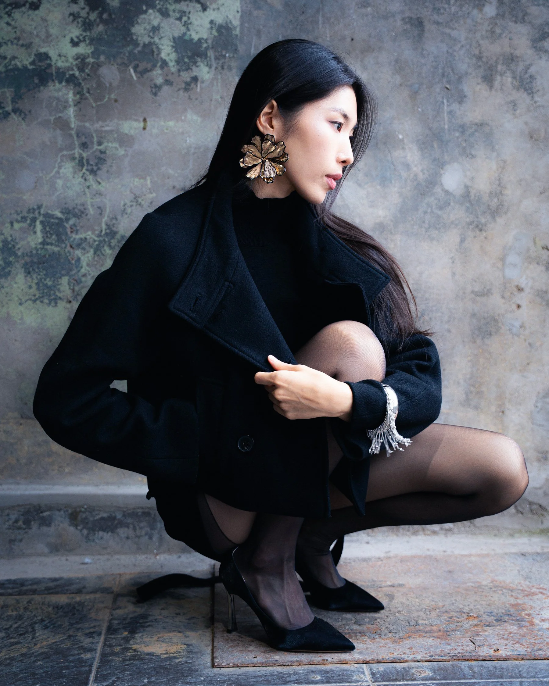 Femme assise en talons hauts noirs, portant un manteau noir, des collants noirs et de grosses boucles d'oreilles en forme de papillon doré. Elle est près d'un mur en béton.