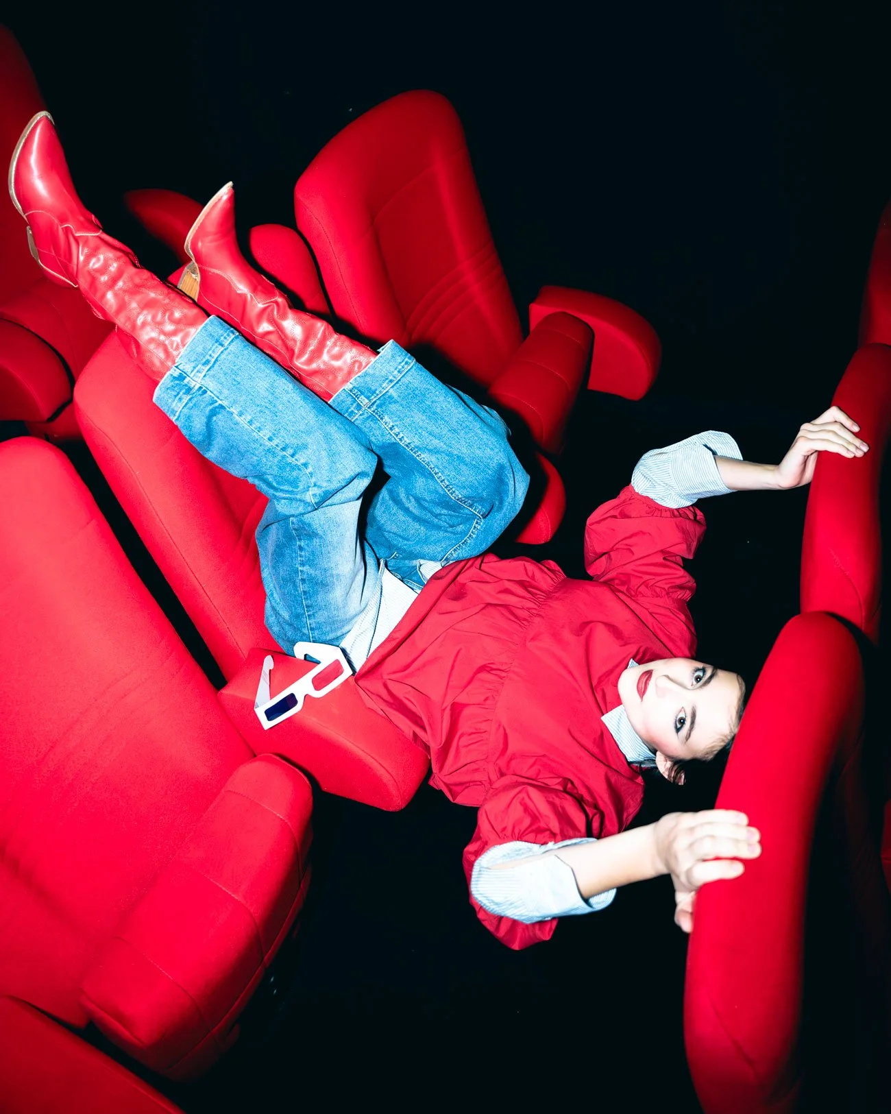 Une femme jeune portant un manteau rouge, des jeans et des bottes rouges, allongée sur des sièges de cinéma rouges, regardant vers l'objectif.