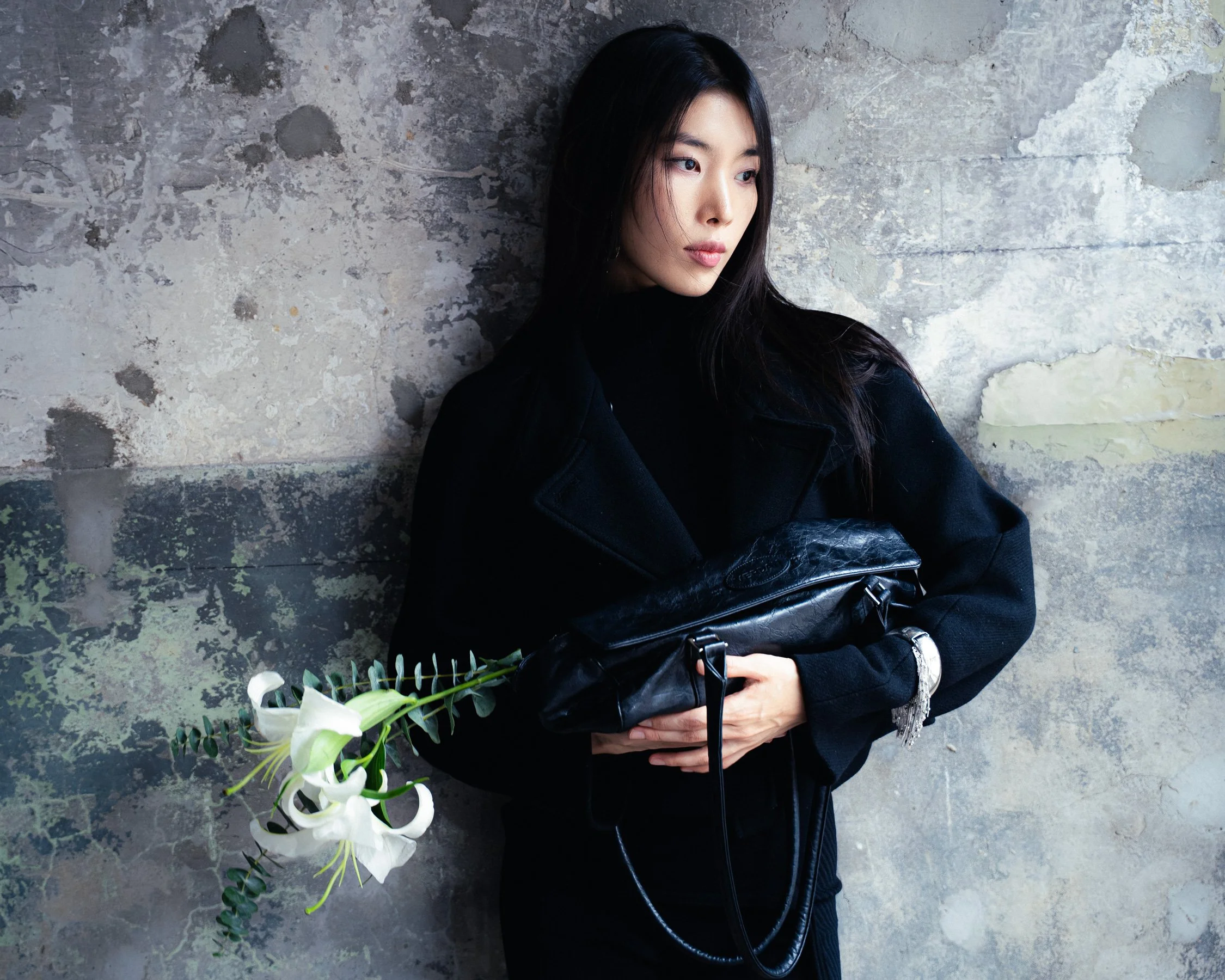 Une femme avec des cheveux longs et noirs, portant un manteau noir, tient un sac noir en caoutchouc, et se tient contre un mur en béton usé avec des fleurs blanches à côté d'elle.