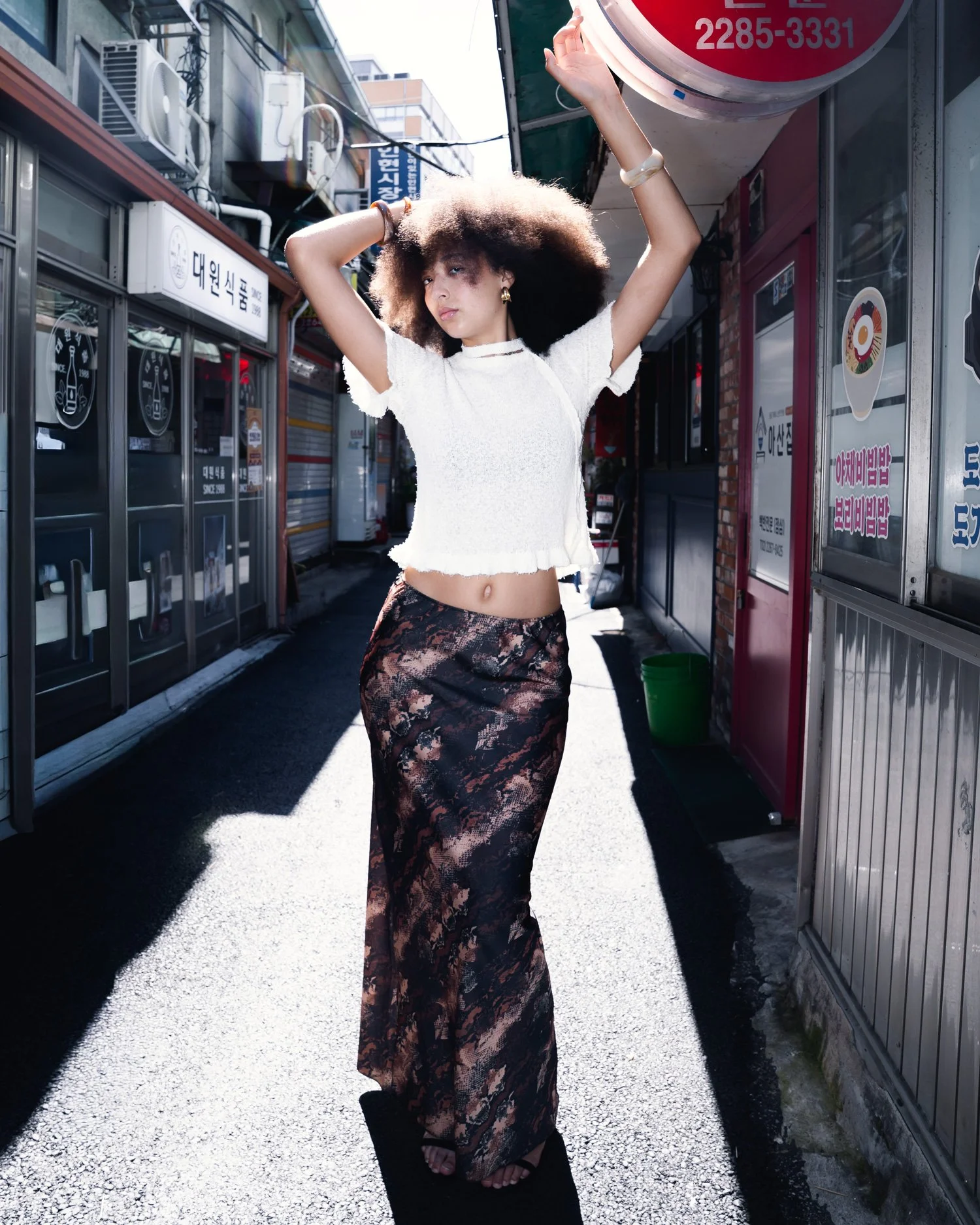 Jeune femme aux cheveux bouclés et afro, portant un crop top blanc et une longue jupe en tissu à motifs, en marchant dans une rue urbaine ensoleillée.