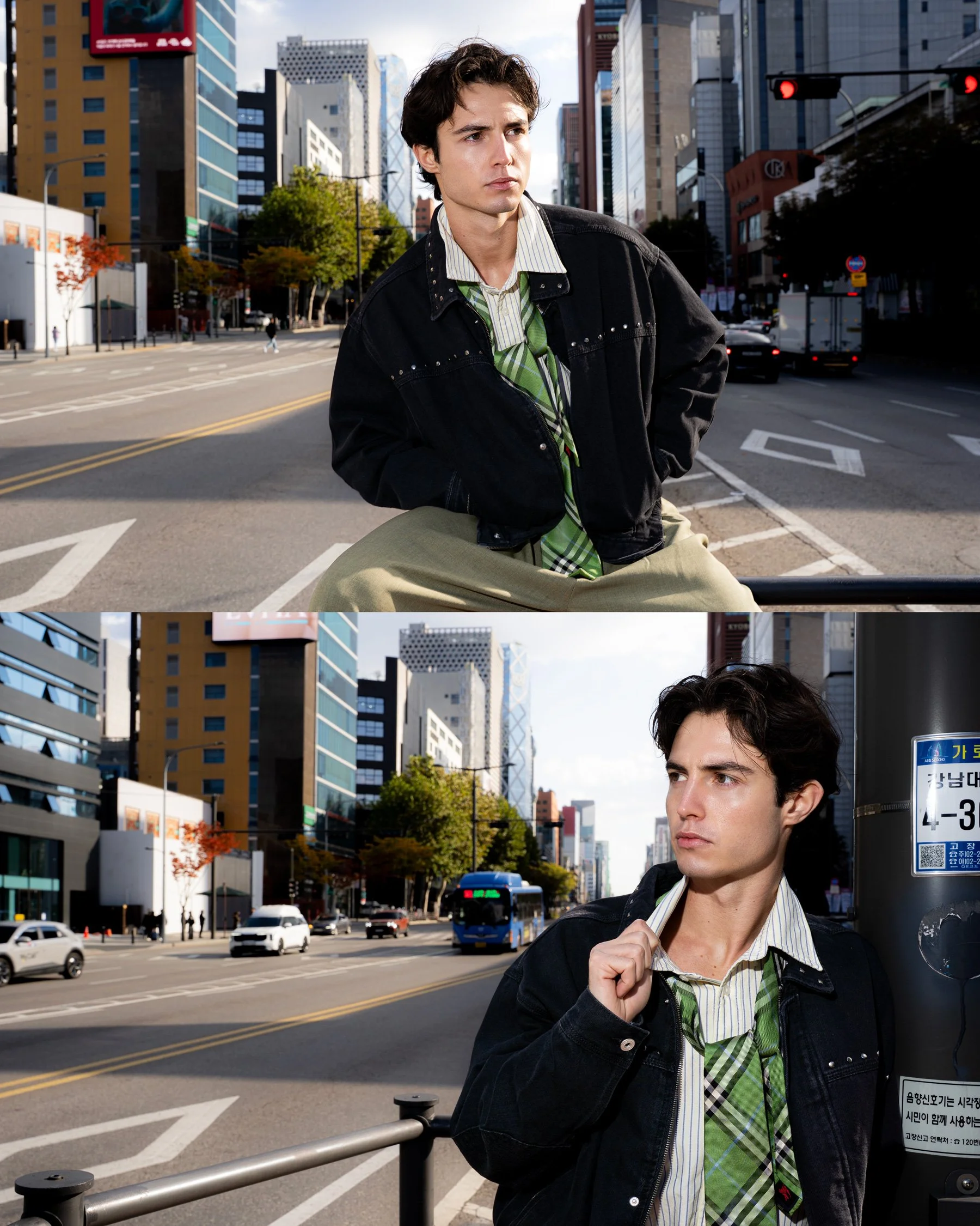 Un jeune homme avec des cheveux noirs, portant une veste noire, une chemise rayée et une cravate à carreaux verts, dans une rue urbaine avec des bâtiments modernes, des voitures et un bus, posant contre un mur et un poteau.