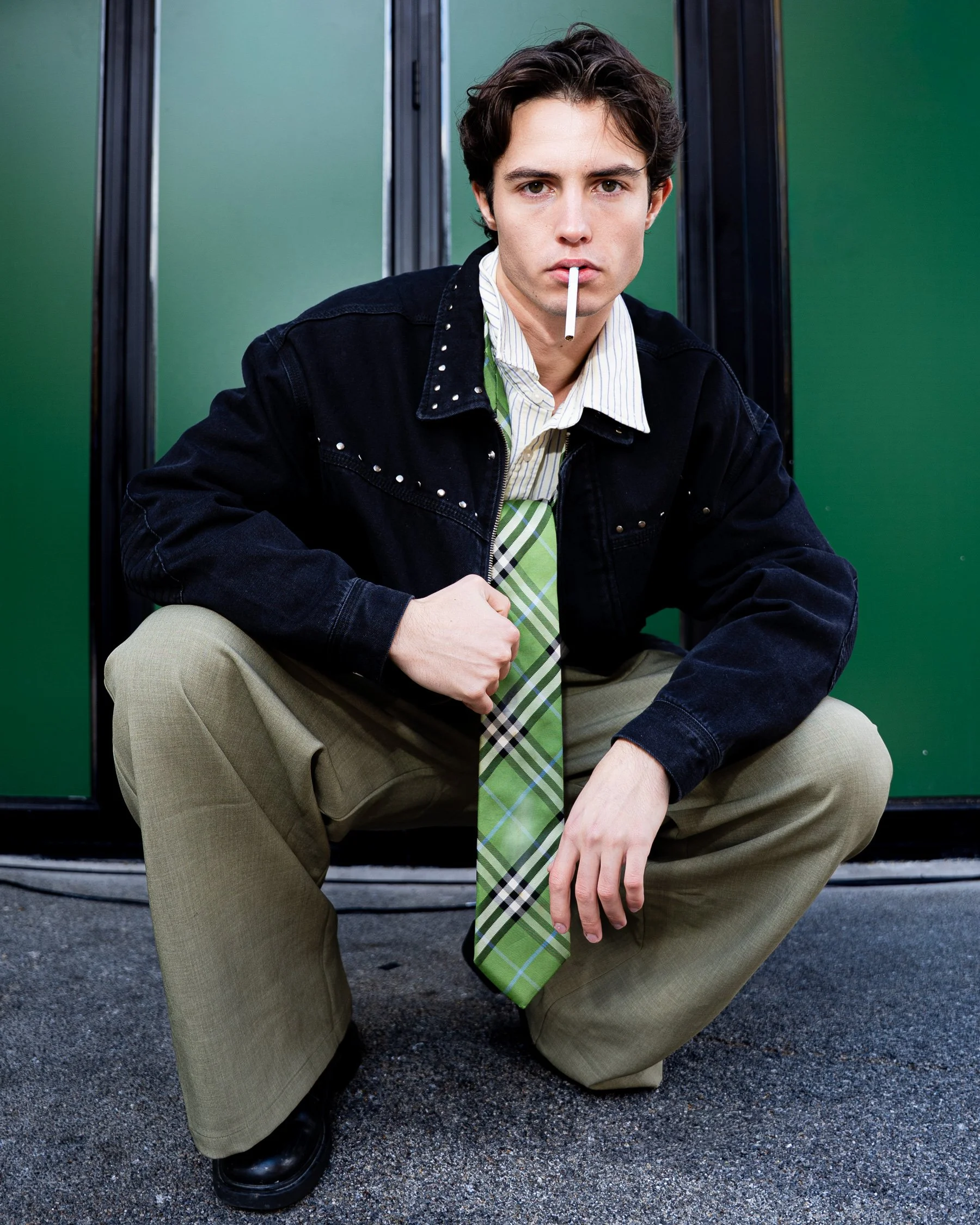 Jeune homme aux cheveux bruns, portant une veste en jean noire, une chemise blanche avec rayures, une cravate à carreaux verts, en position accroupie devant un fond vert.