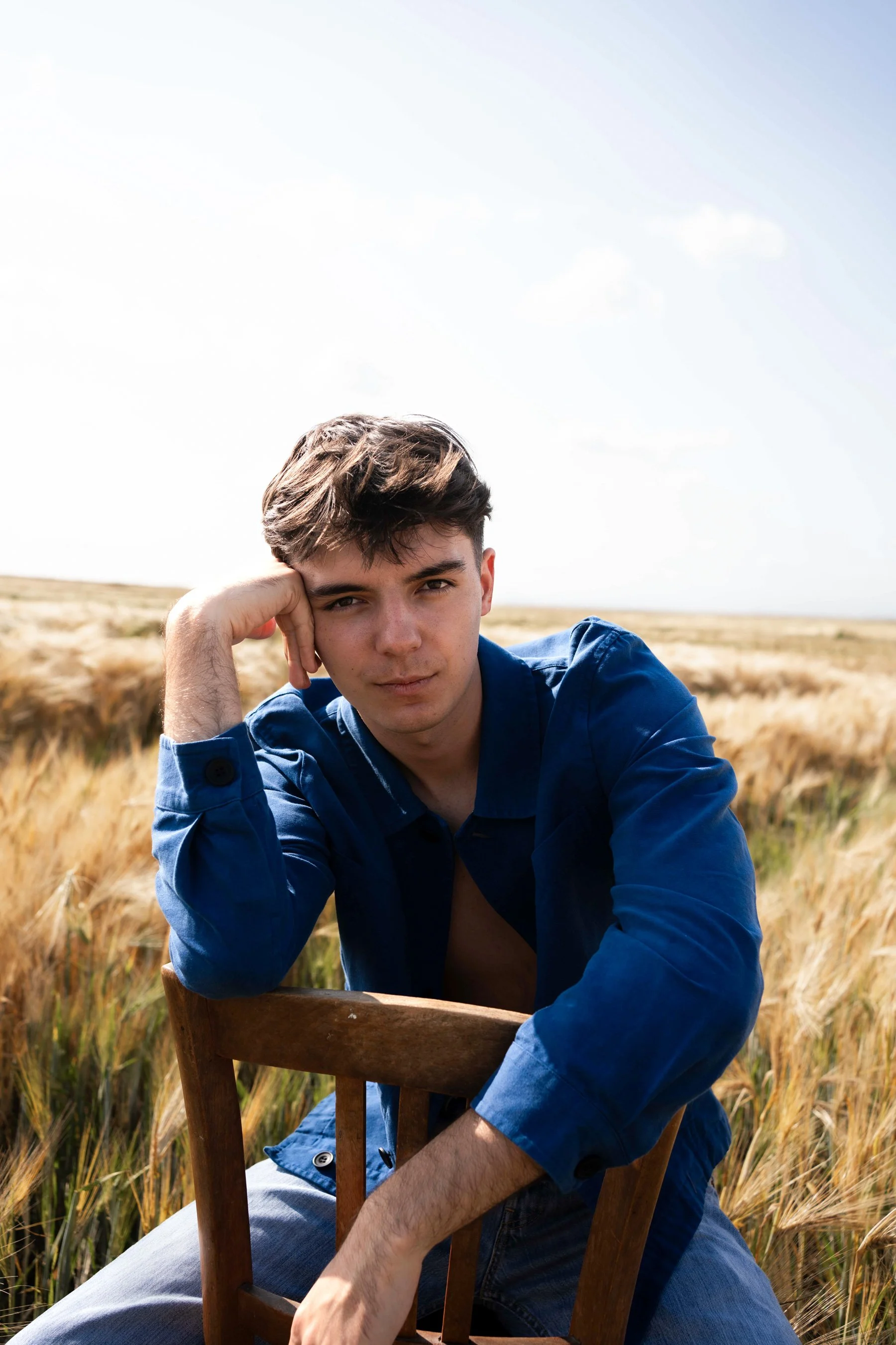 Jeune homme assis sur une chaise en bois dans un champ de blé, regardant l'objectif, portant une chemise bleue.