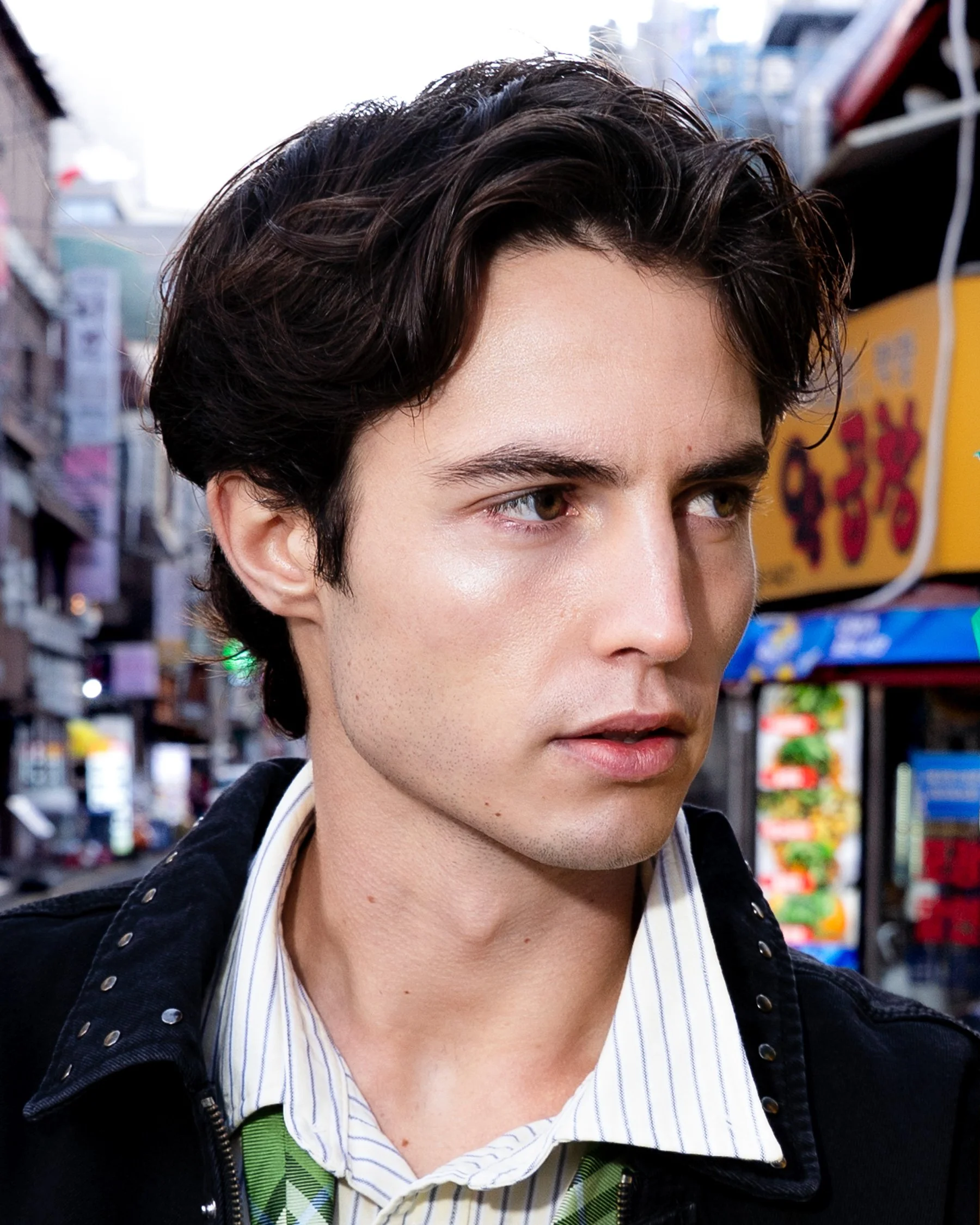 Un jeune homme avec des cheveux bruns et ondulés, portant une chemise à rayures et une veste noire, se promène dans une rue urbaine animée.