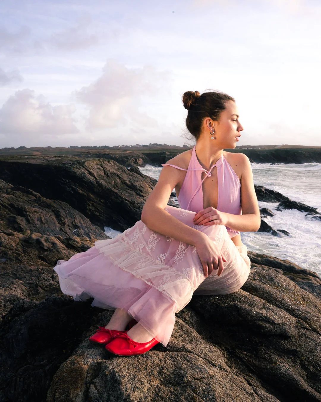 Jeune femme assise sur des rochers à la plage, portant une robe rose, avec des cheveux en chignon, regardant à droite, avec l'océan et le ciel nuageux en arrière-plan.