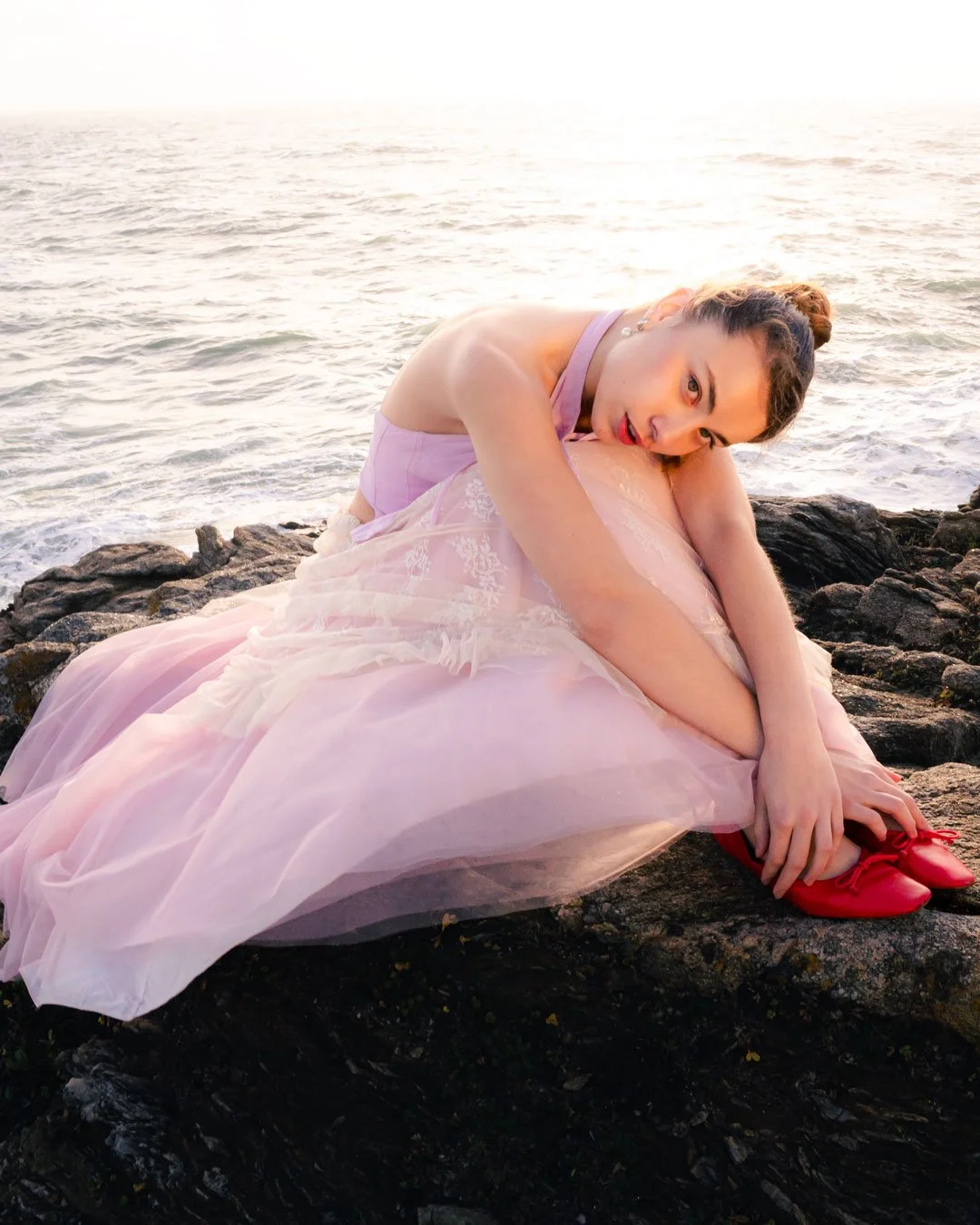 Une femme en robe rose posant sur des rochers près de la mer au coucher du soleil.