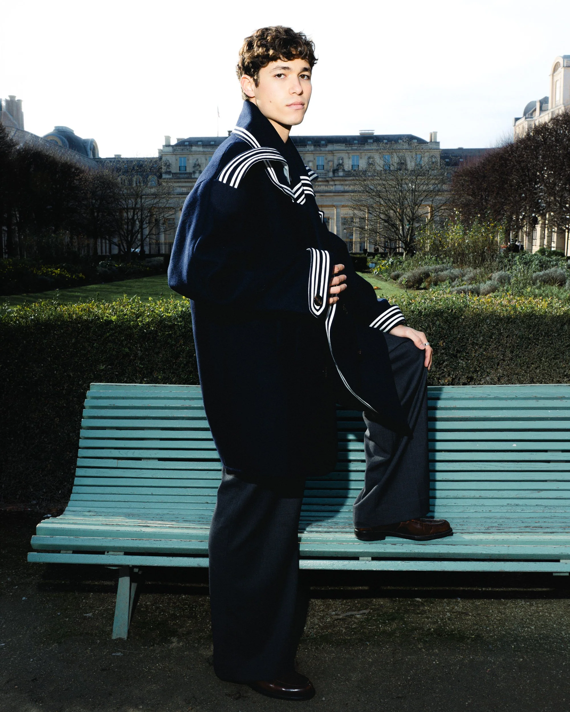 Un jeune homme pose sur un banc dans un parc, portant une veste noire avec des détails en blanc et une chemise, avec un bâtiment classique en arrière-plan.