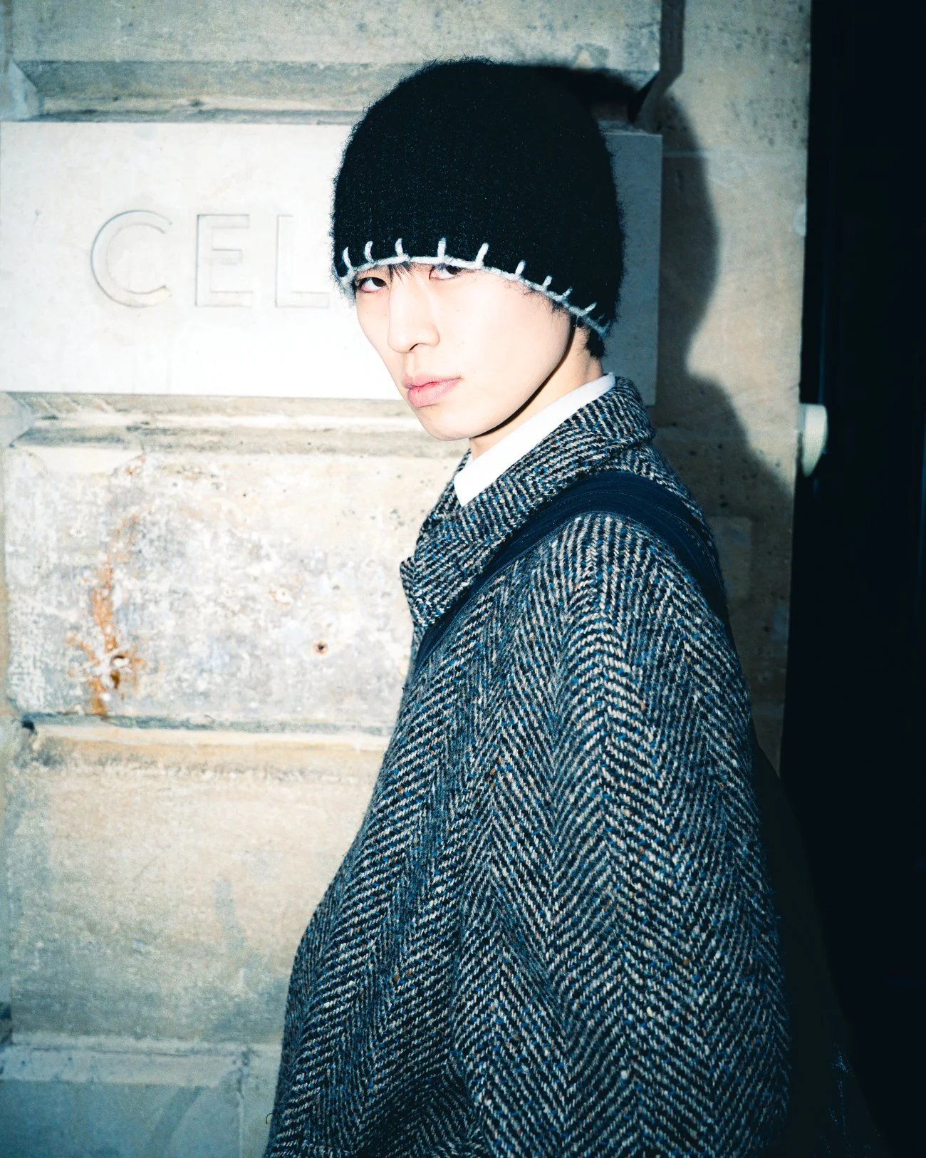 Jeune homme portant un bonnet noir à bord blanc, veste en tweed, en face d'un mur en pierre gravé avec le mot 'CELINE'.
