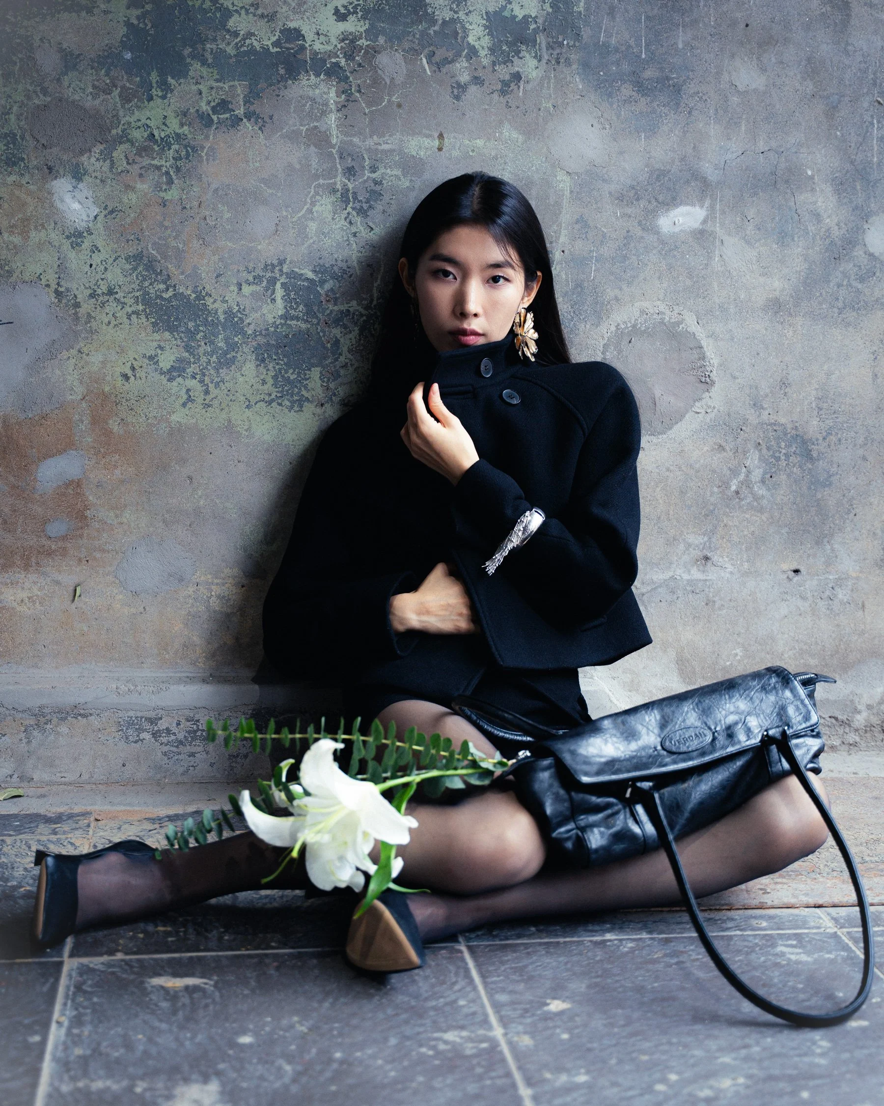 Une femme assise contre un mur en pierre, portant un manteau noir, avec un sac en cuir noir à ses côtés et des fleurs blanches sur ses genoux.