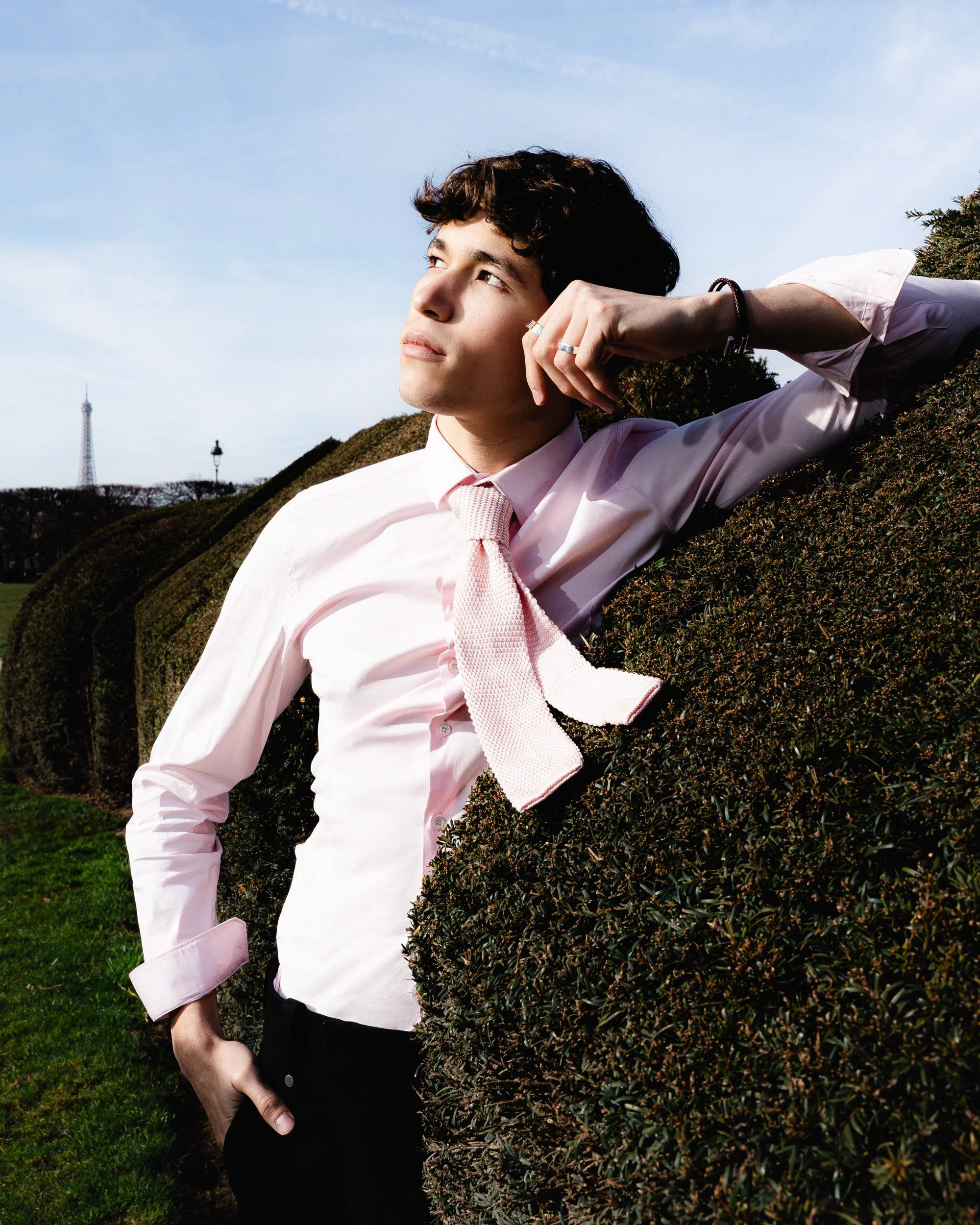 Jeune homme en chemise rose et cravate, posant près d'une haie, regardant vers le ciel, avec la Tour Eiffel en arrière-plan.