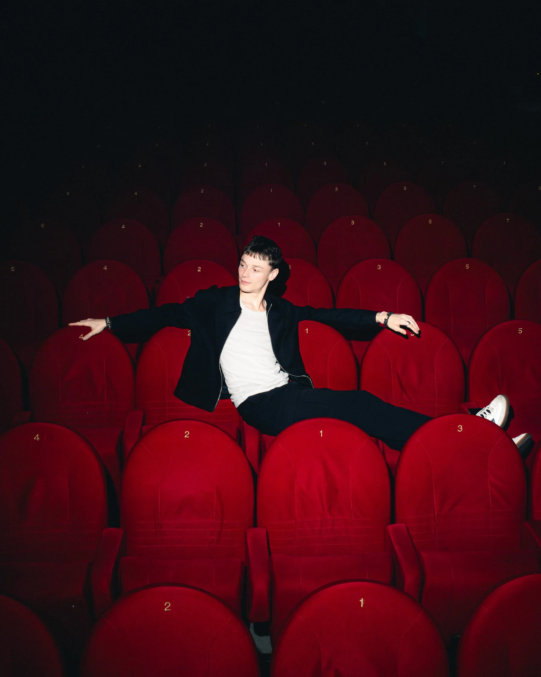 Une personne assise dans une salle de théâtre ou cinéma, entourée de fauteuils rouges, portant une veste noire et un t-shirt blanc, en position décontractée.