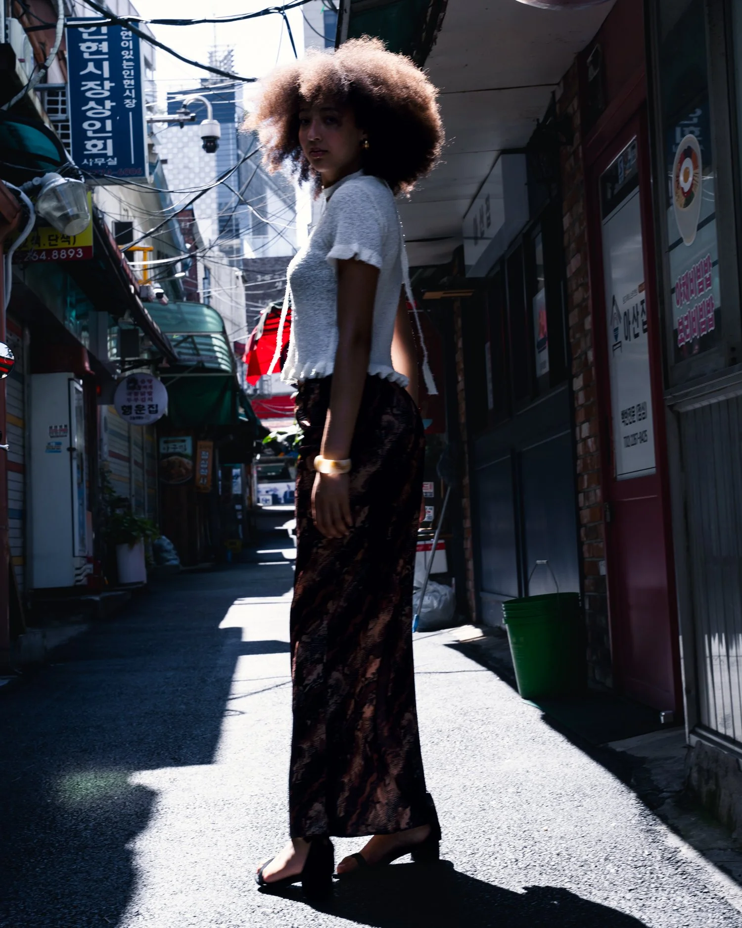 Une jeune femme marche dans une ruelle étroite, portant une coiffure volumineuse et un vêtement léger. La scène est en plein jour avec des panneaux en coréen et divers stands ou magasins sur le côté.