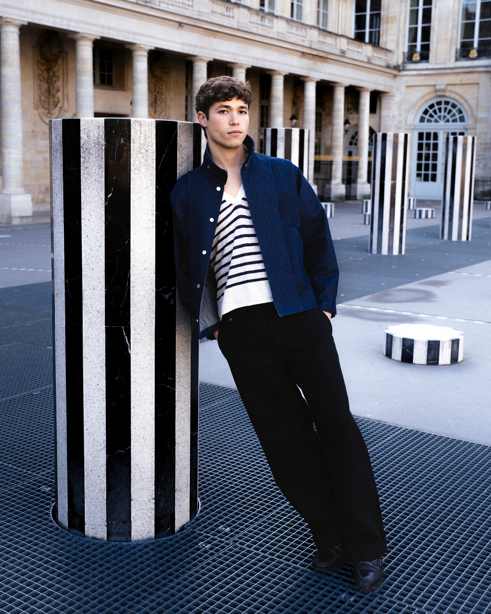 Jeune homme debout près d'une colonne noire et blanche dans une place publique historique, construite en pierre avec des colonnes et des fenêtres en arc.