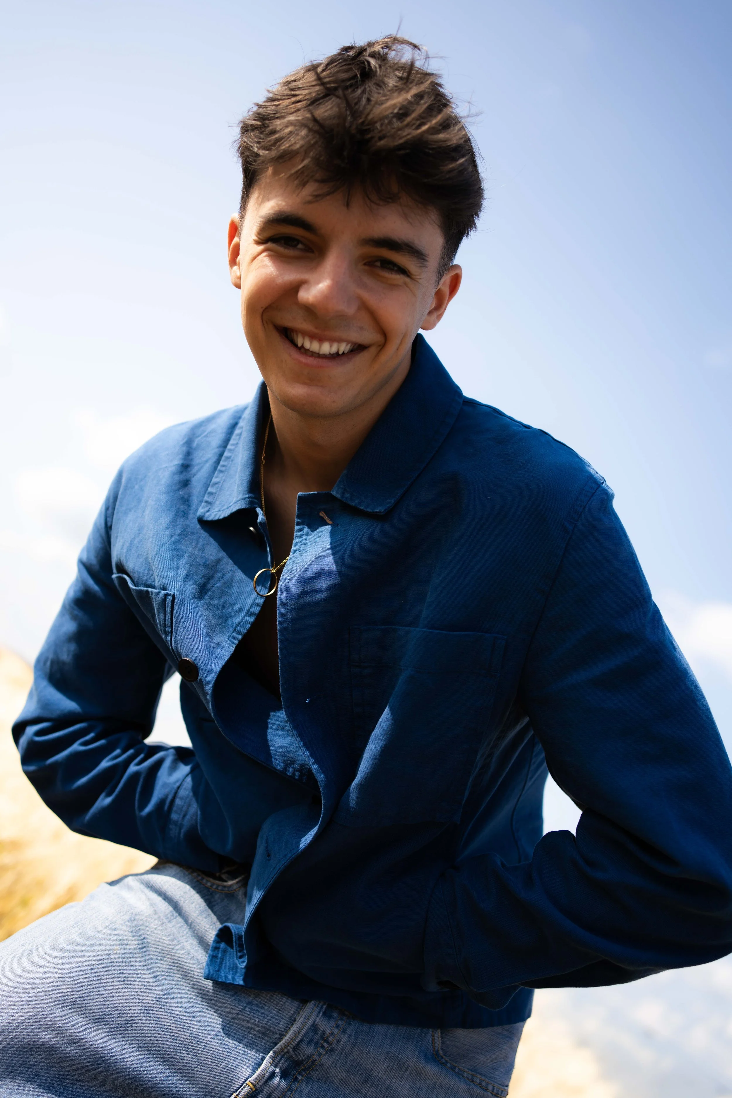 Jeune homme souriant portant une veste bleue et un jean, debout à l'extérieur avec un ciel bleu en arrière-plan.