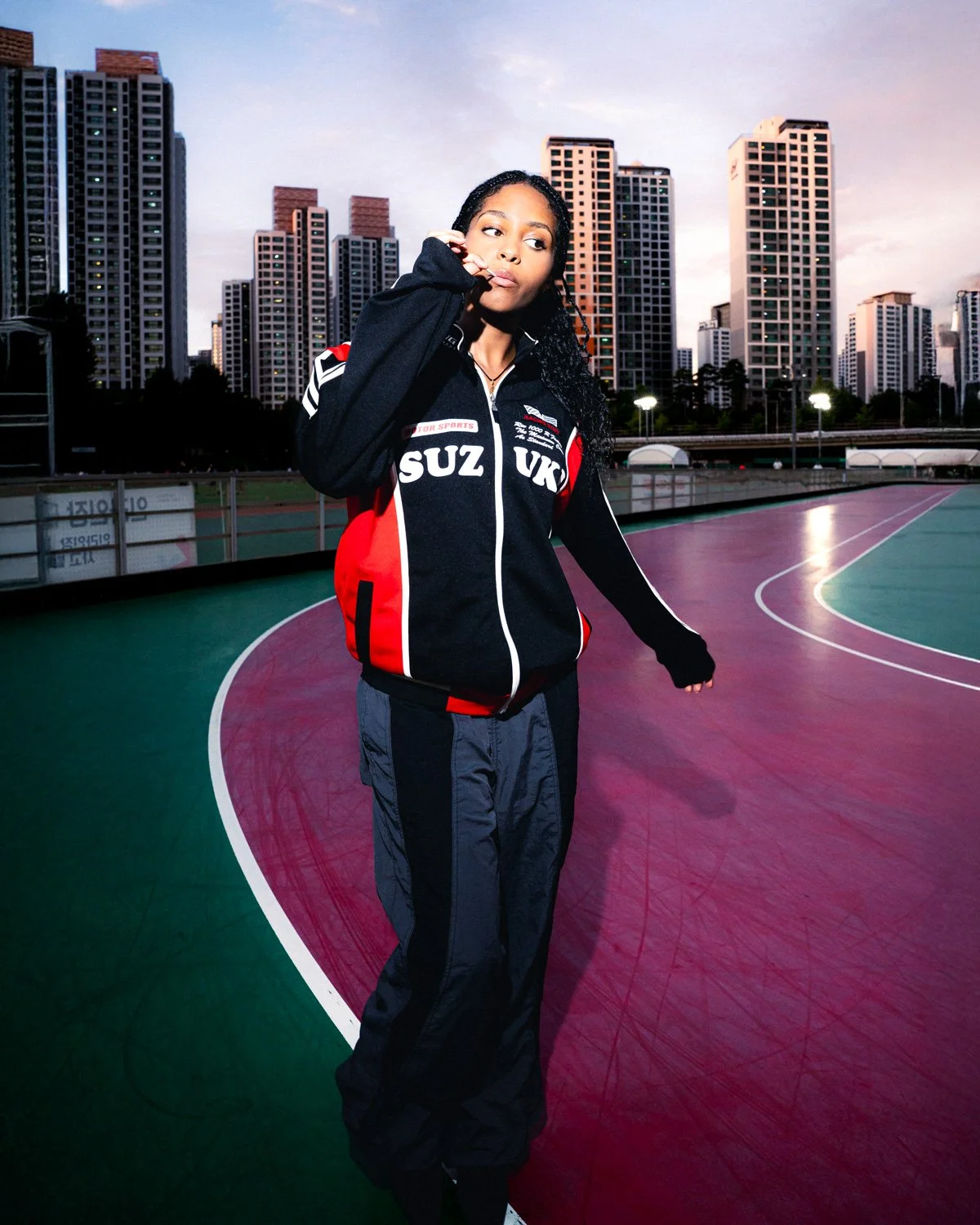 Une jeune femme avec des cheveux bouclés portant un sweat à capuche noir avec des détails rouges et blancs, et un pantalon noir, se tient sur une piste de sport en ville au crépuscule, avec des gratte-ciel en arrière-plan.