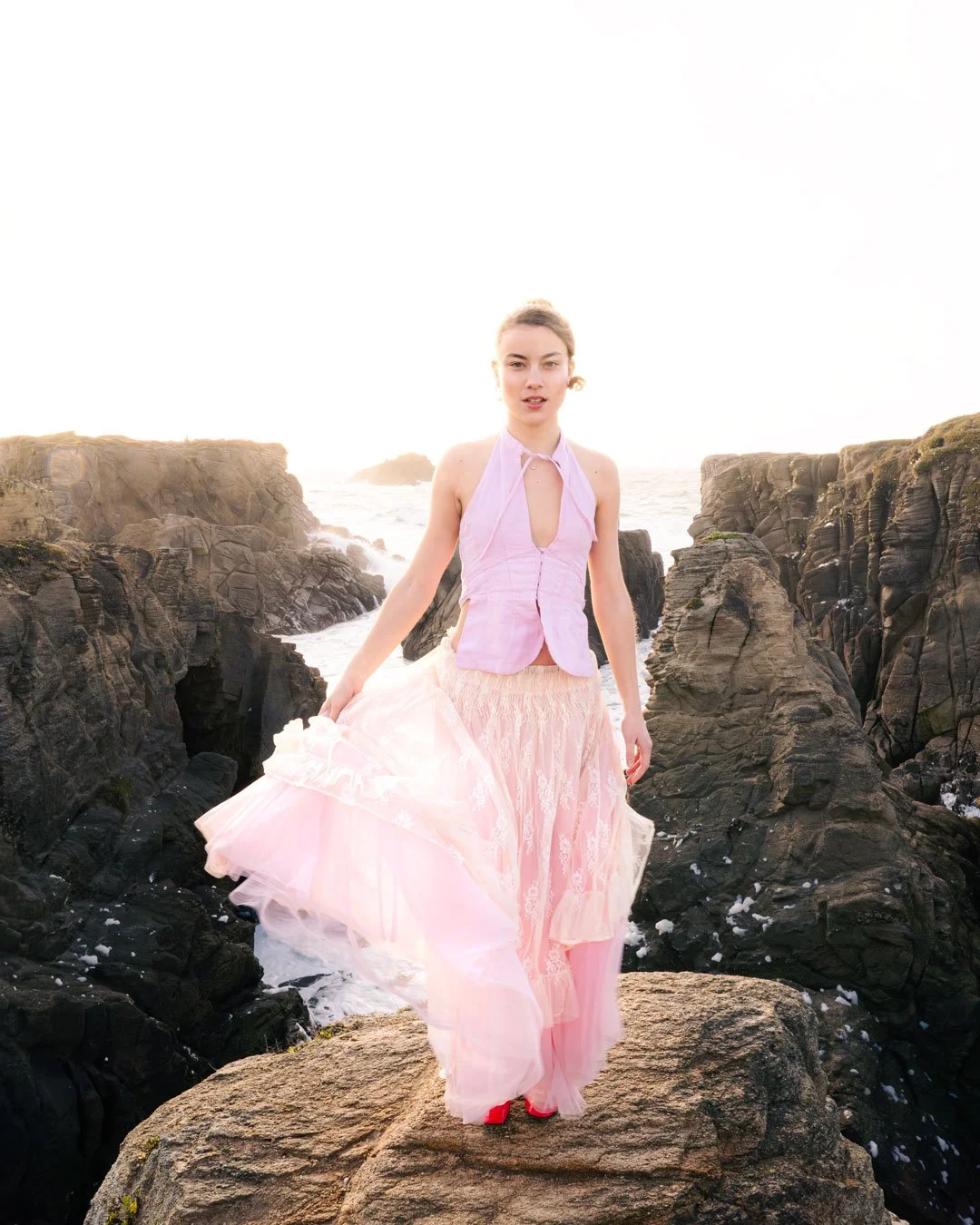 Femme en robe rose claire sur une roche près de la mer, avec des falaises rocheuses en arrière-plan, au coucher du soleil.