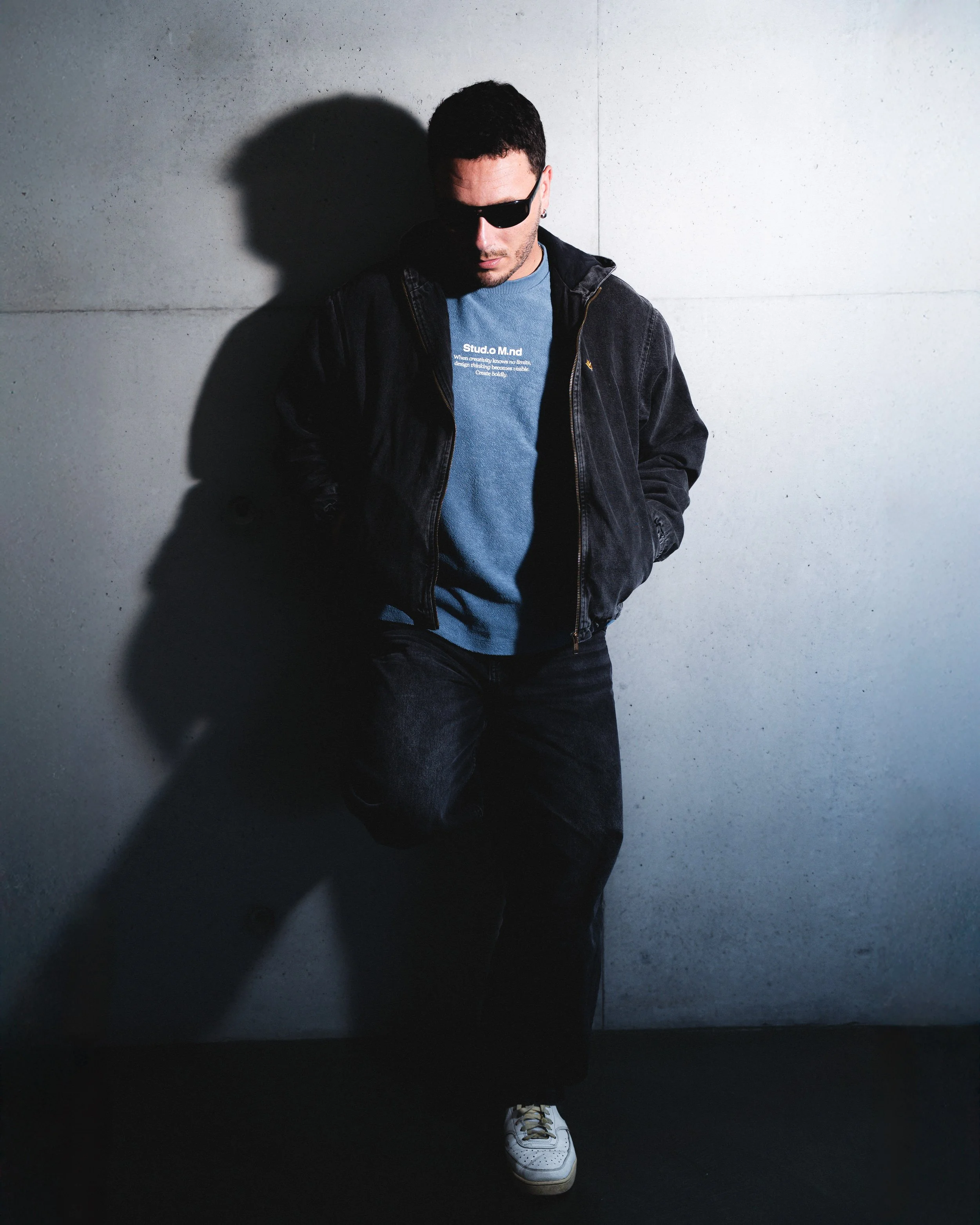 Jeune homme portant des vêtements noirs et une chemise bleue, avec des lunettes de soleil, appuyé contre un mur en béton.