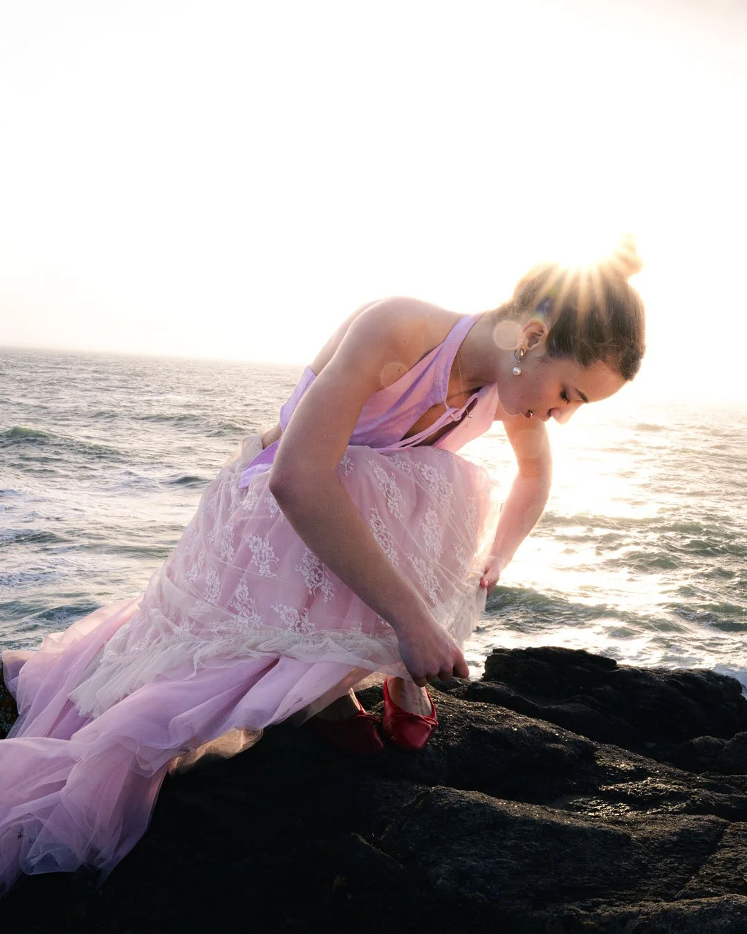 Une femme en robe rose et chaussures rouges, accroupie sur des rochers près de la mer, au coucher du soleil.