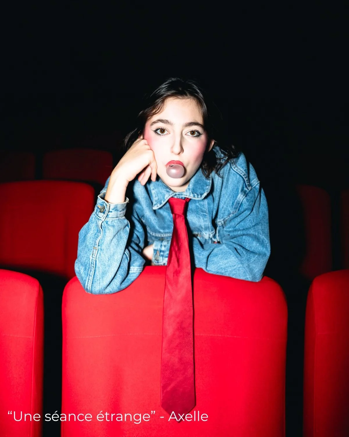 Une jeune femme assise dans une salle de cinéma avec des sièges rouges, portant une veste en jean et une cravate rouge, avec du maquillage rouge autour des yeux et des joues, et une boule de gomme dans la bouche.