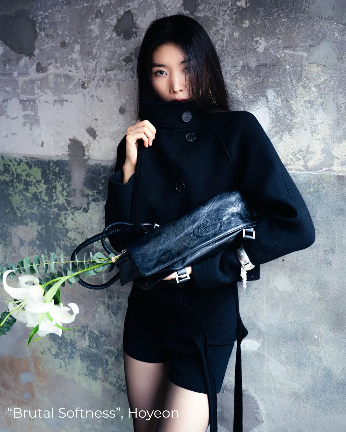 Une jeune femme avec des cheveux noirs portant un manteau noir et un sac noir, se tenant devant un mur en pierre, tenant une branche de fleurs blanches.