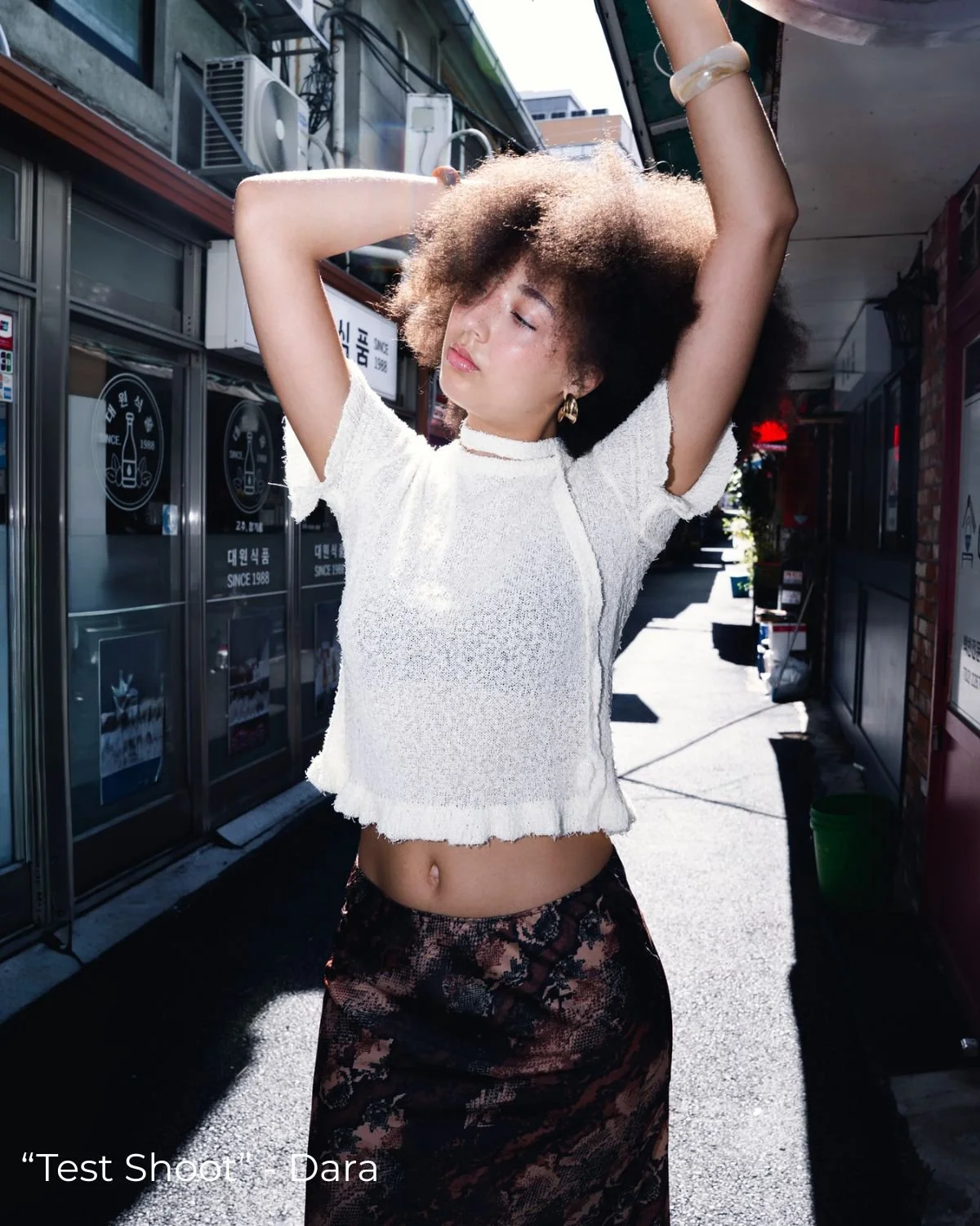 Jeune femme fixant vers le bas, bras levés, portant un haut blanc en tissu léger et une jupe longue à motifs. Elle se trouve dans une rue ensoleillée avec des bâtiments en arrière-plan.