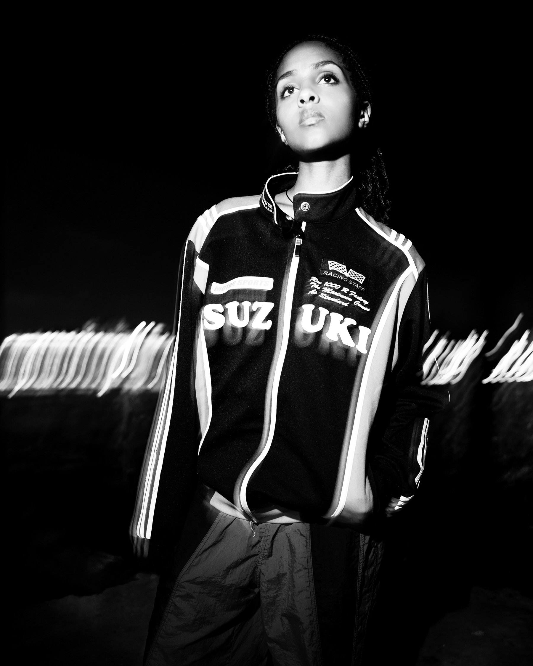 Jeune femme noire posant sous un éclairage nocturne, portant une veste de course UKI avec des logos, en noir et blanc.
