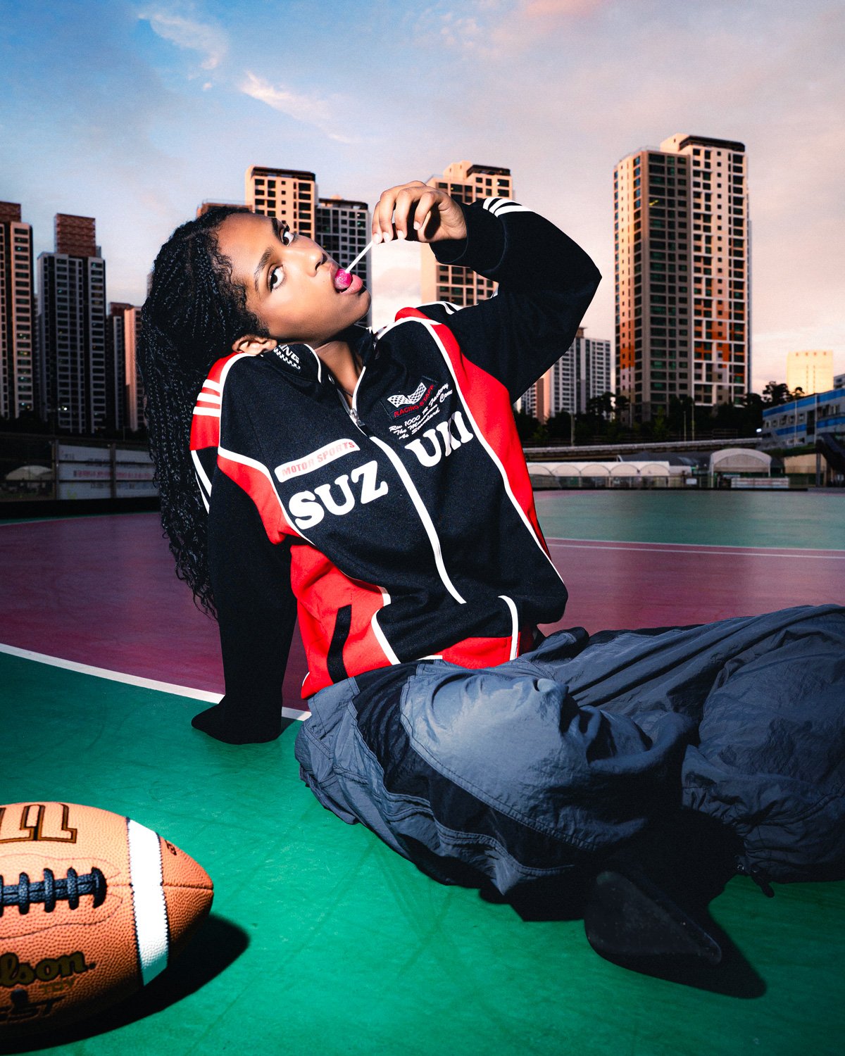 Jeune femme rousse portant une veste de sport noire et rouge, assise sur un terrain de basketball extérieur avec des immeubles en arrière-plan, en train de manger une sucette.