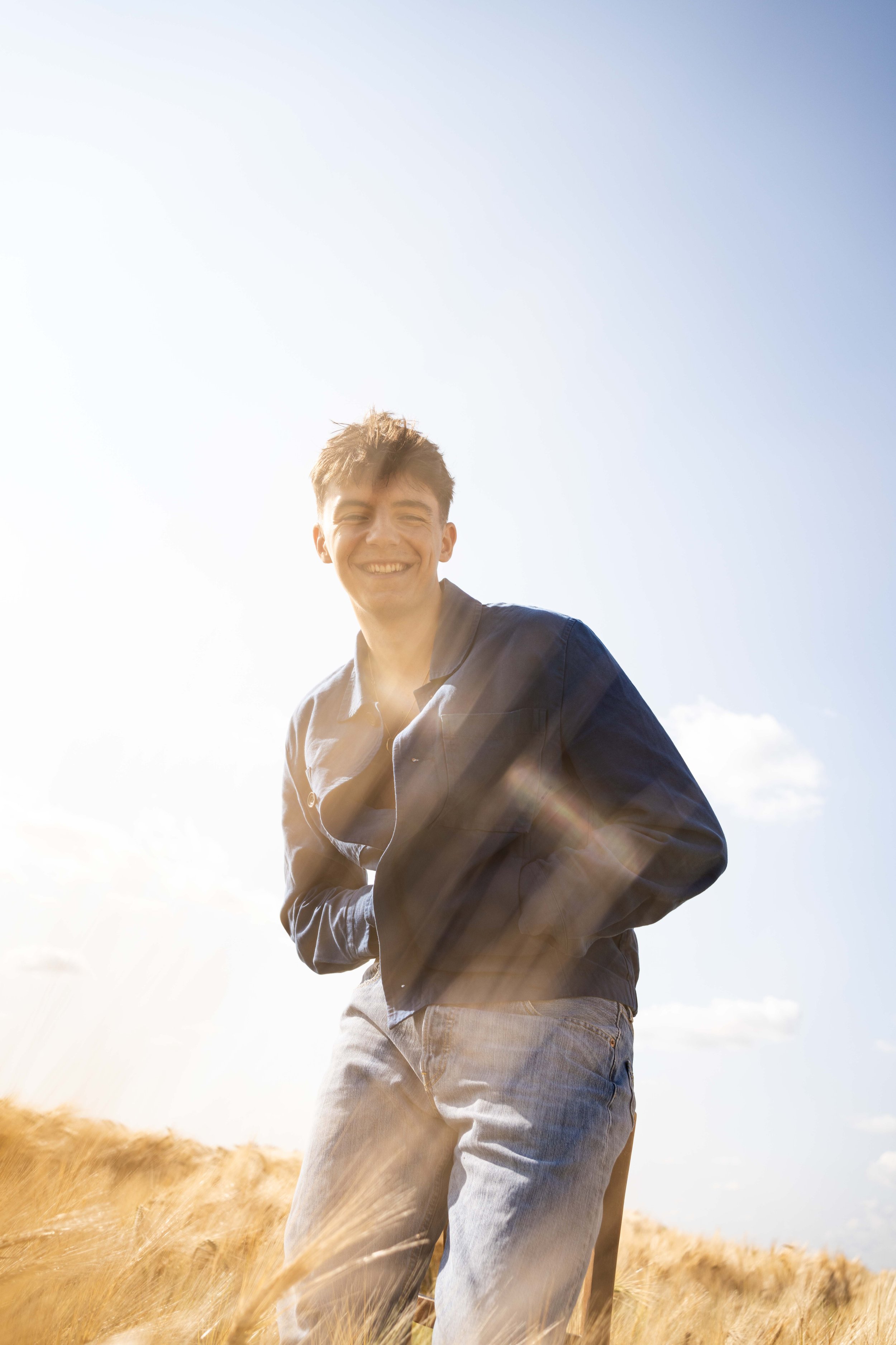 Jeune homme souriant debout dans un champ de blé sous un ciel ensoleillé.