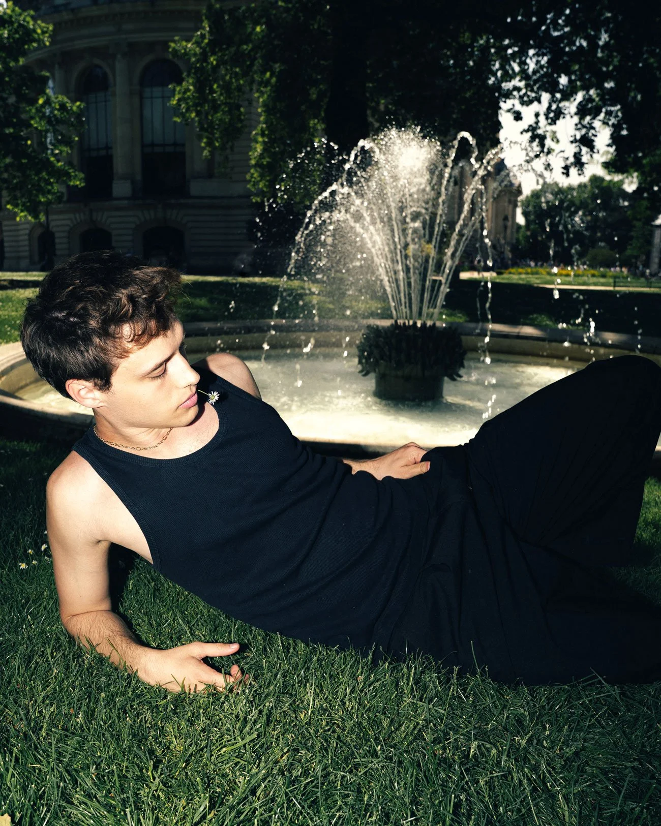 Jeune homme allongé sur l'herbe devant une fontaine en plein air, entouré d'arbres et d'un bâtiment historique en arrière-plan.