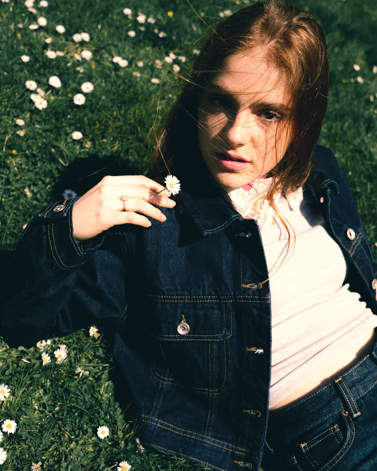 jeune femme avec cheveux longs, portant un jean, allongée dans un champ de marguerites, souriant légèrement