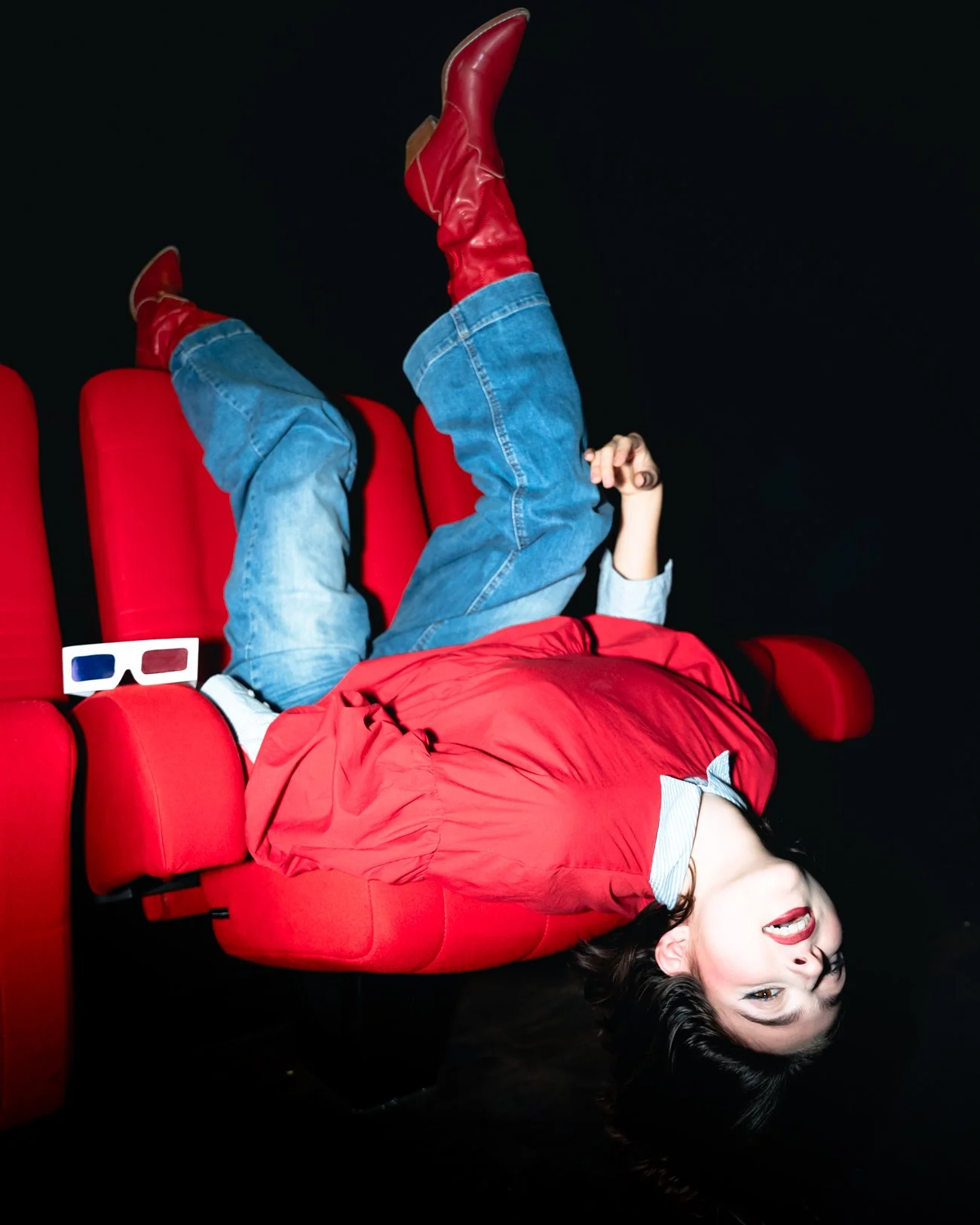 Une femme souriante allongée sur un fauteuil rouge, portant un blouson rouge, un jean et des bottes rouges, avec des lunettes 3D posées à côté d'elle.