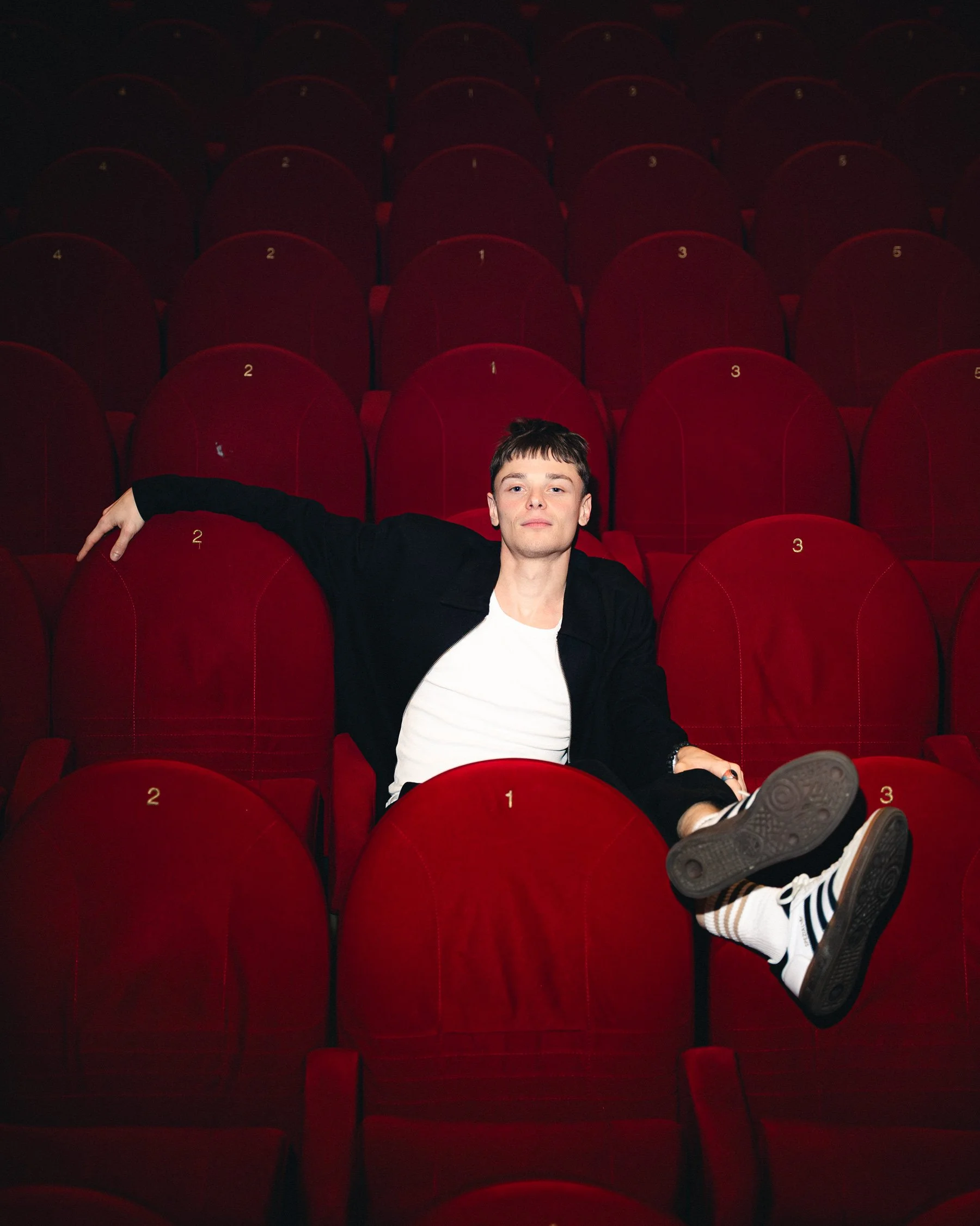 Un jeune homme assis dans une salle de cinéma avec des sièges rouges, portant un t-shirt blanc, un blazer noir, et des sneakers, en position décontractée.