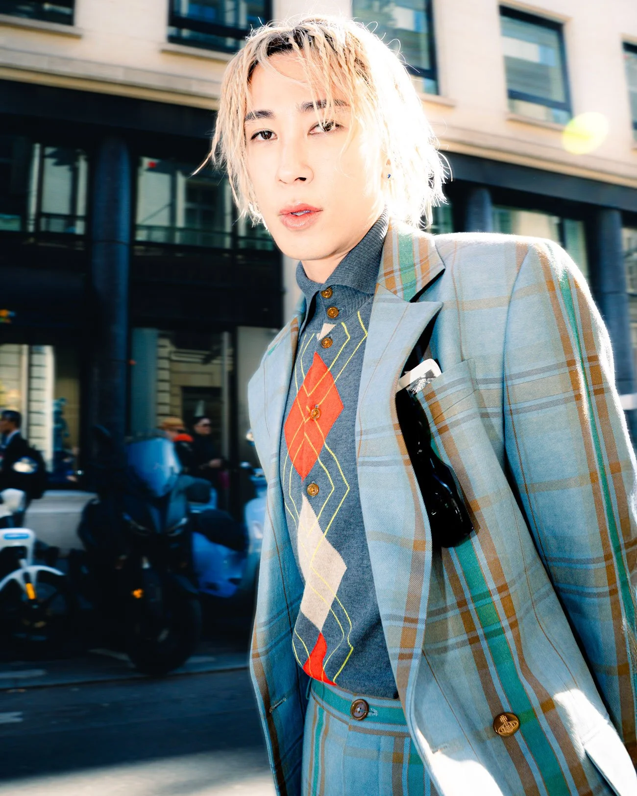 Jeune homme à cheveux blonds et bouclés, portant un costume à carreaux, dans une rue urbaine.