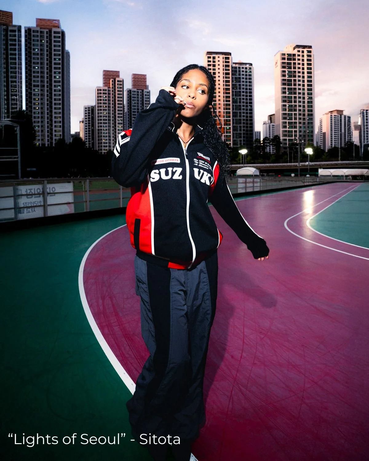 Une femme debout sur une piste d'athlétisme avec une ville moderne en arrière-plan, portant une veste noire avec des éléments rouges et blancs.
