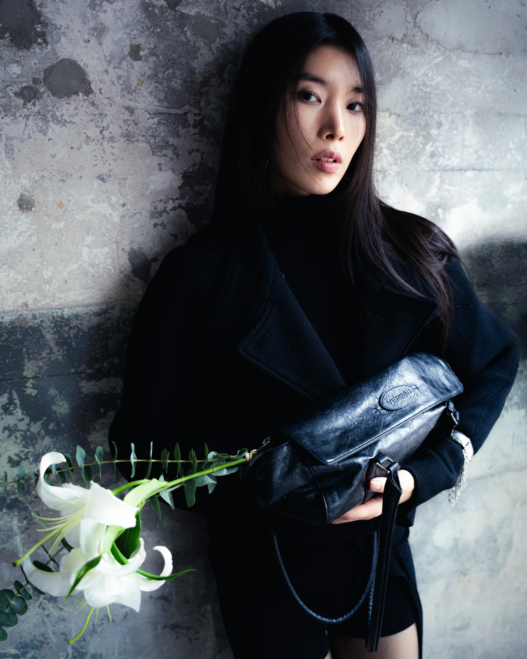 Jeune femme asiatique avec des cheveux longs, portant un manteau noir et tenant un sac en cuir noir, posant contre un mur en pierre avec des fleurs blanches à côté.