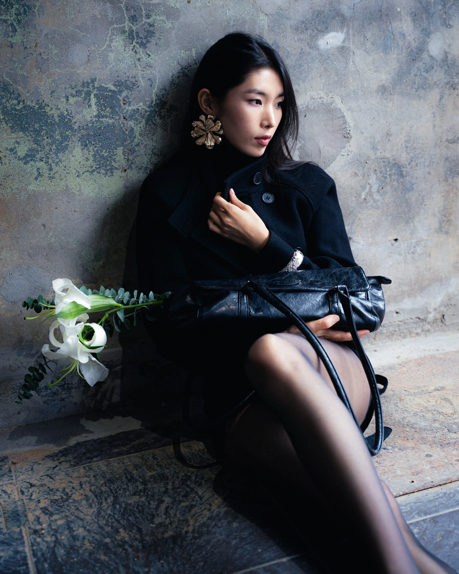 Une femme assise contre un mur en plâtre à l'aspect vintage, portant un manteau noir, des collants transparents, des boucles d'oreilles volumineuses en forme de fleurs, et tenant un sac en cuir noir, avec un bouquet de fleurs blanches à ses côtés.