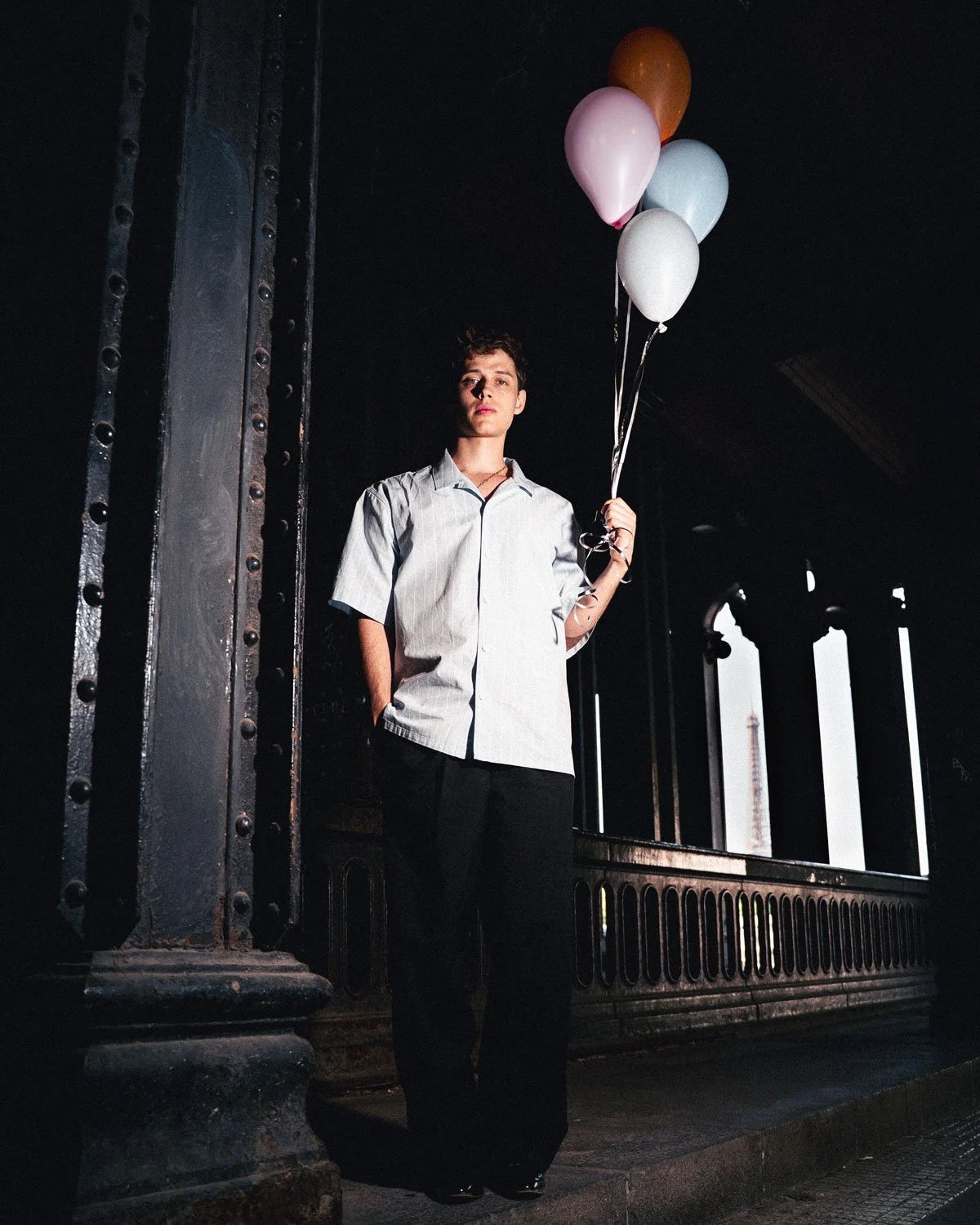 Jeune homme debout sous un pont, tenant un bouquet de ballons colorés, portant une chemise blanche et un pantalon noir, dans un environnement urbain en fin d'après-midi
