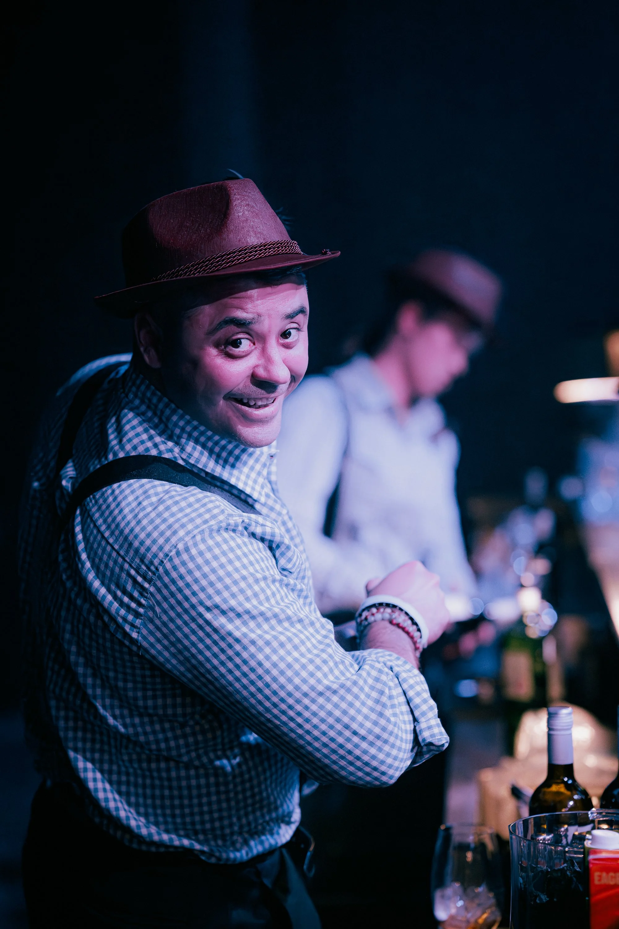 A man smiling and looking at the camera, wearing a brown fedora, checkered shirt, and suspenders at a bar or party, with a woman in the background also wearing a hat.