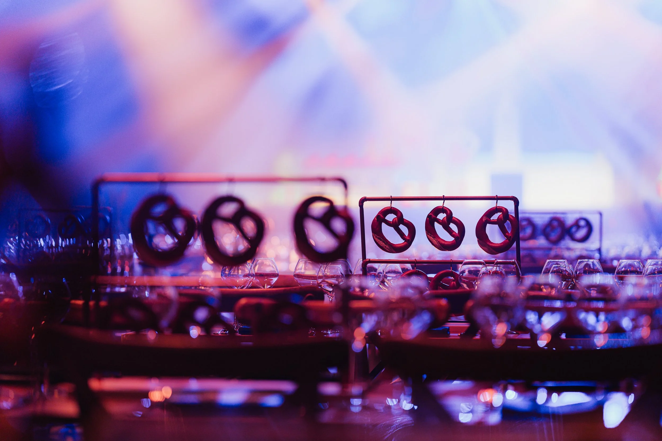 Blurred image of various pretzels, some hanging on racks and some on trays, illuminated in purple and pink lighting.