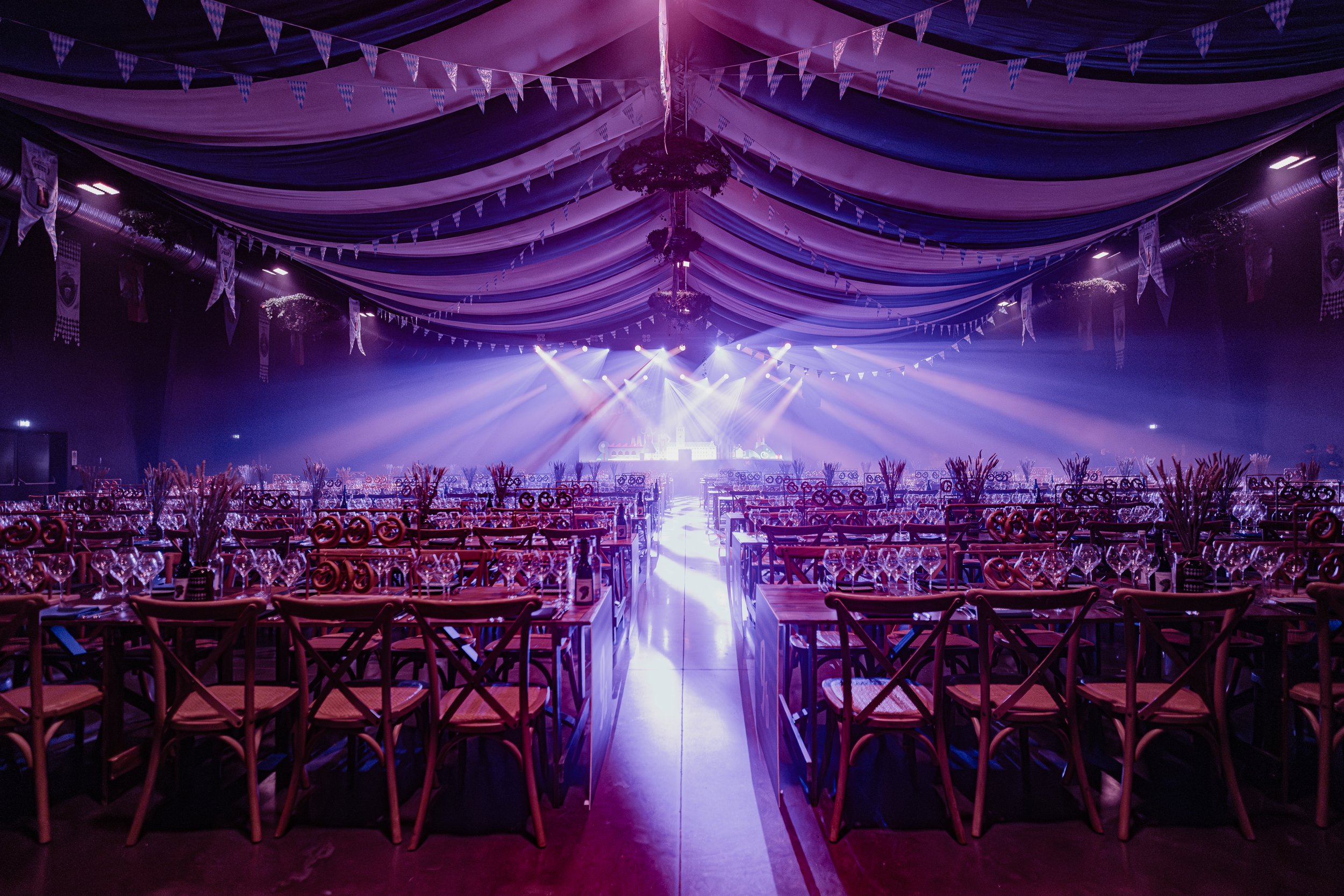 Empty event hall decorated with purple and white bunting hanging from the ceiling, elegant tables and chairs set with wine glasses and centerpieces, illuminated by stage lights at the front of the room.
