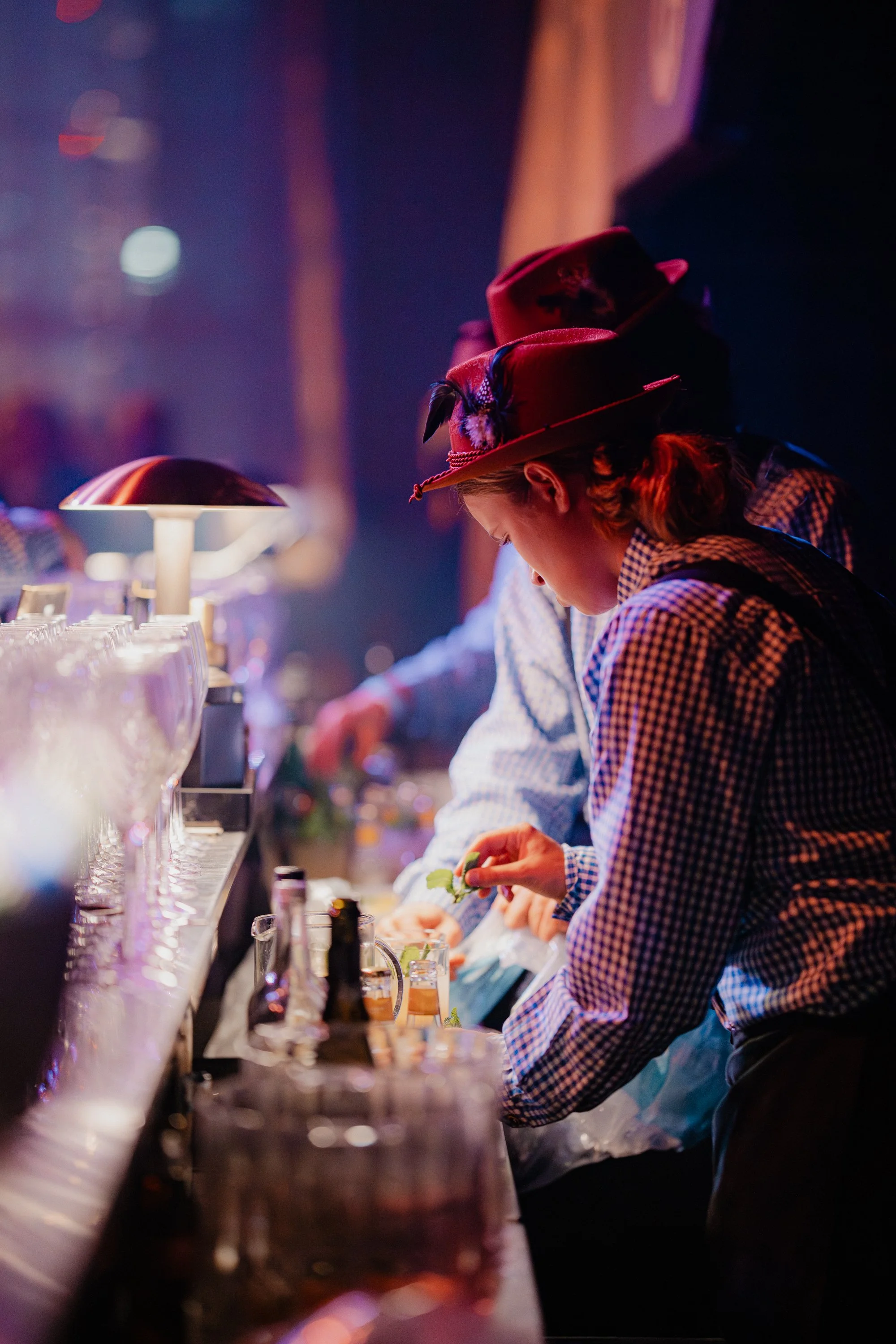 People dressed in traditional Bavarian clothing preparing drinks at a bar, with dim colorful lighting.