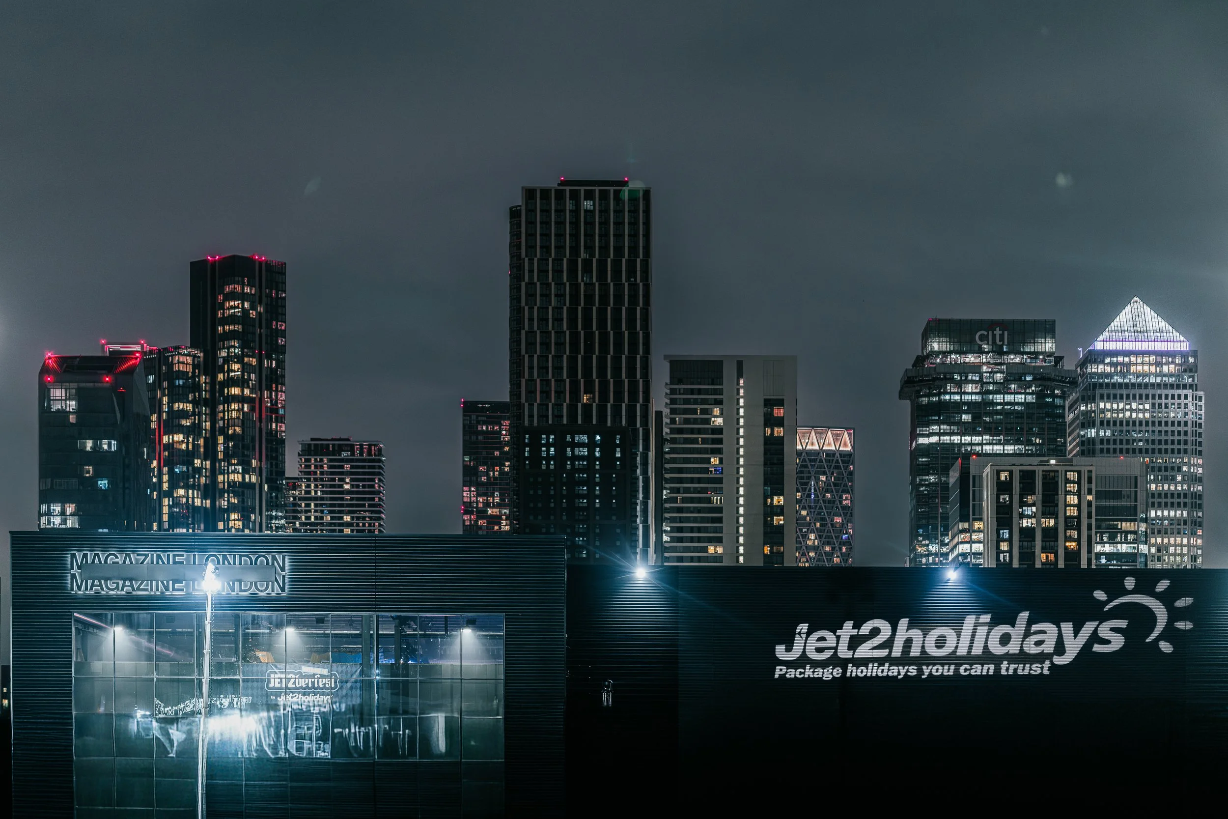 Nighttime cityscape with illuminated skyscrapers and a Jet2holidays building in the foreground.