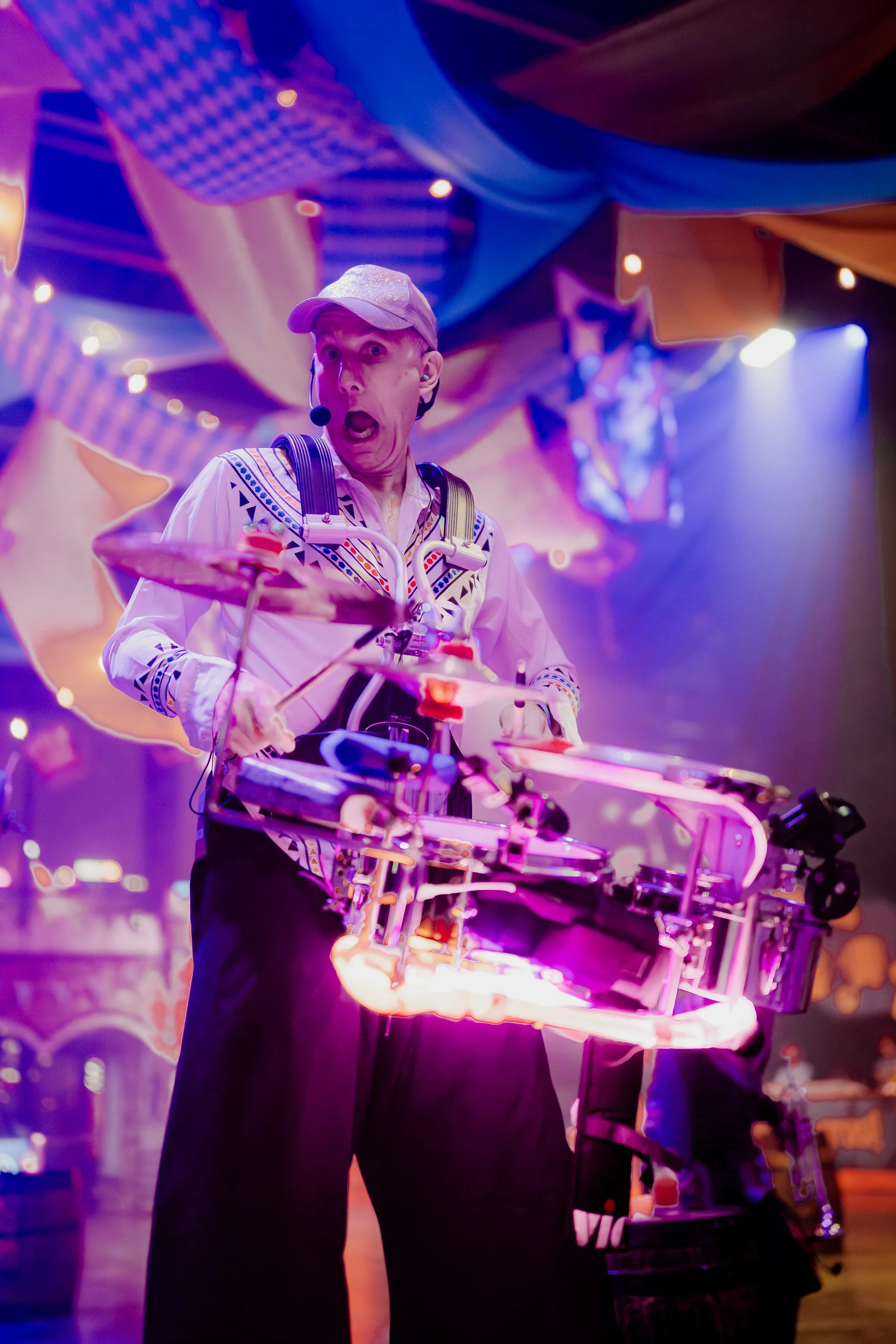 A man performing on stage, wearing a baseball cap, with a headset microphone, holding electronic drum pads, under colorful stage lighting with a vibrant, decorative background.