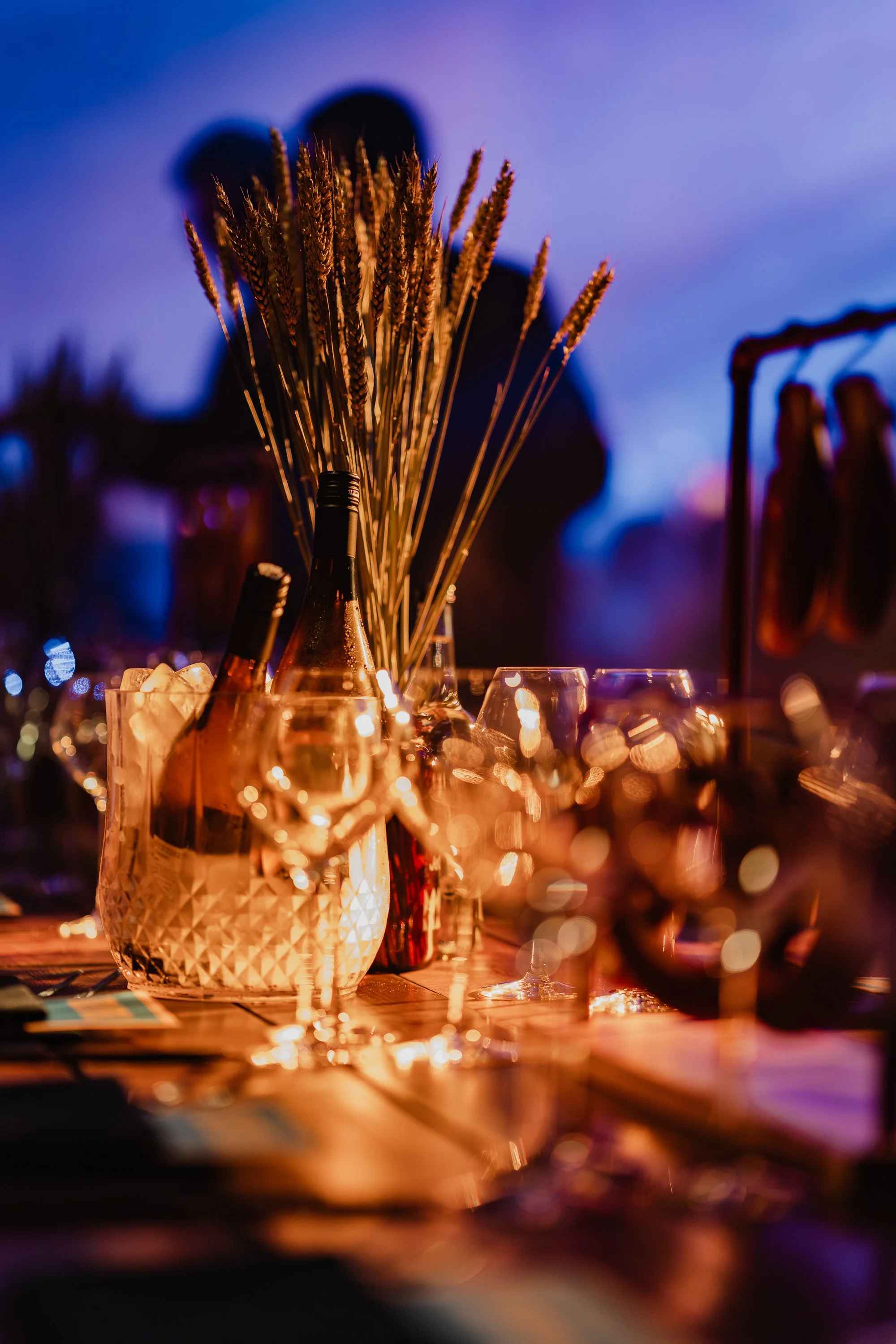 A table with glasses, a bottle, and decorative dried wheat stalks, illuminated by warm lighting at an outdoor evening event.