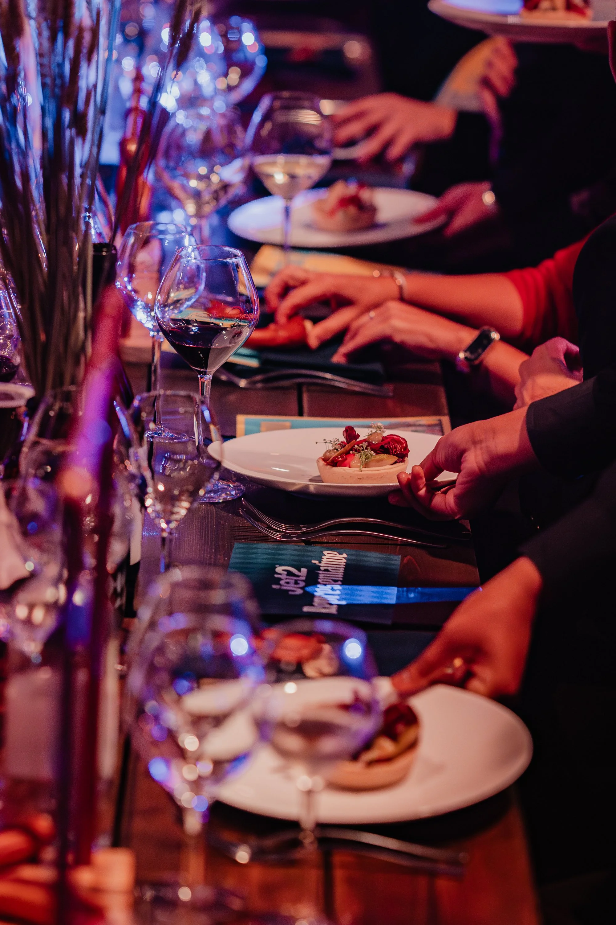 People seated at a long dining table with plates of food and glasses of wine, in a dimly lit setting with purple and red lighting.