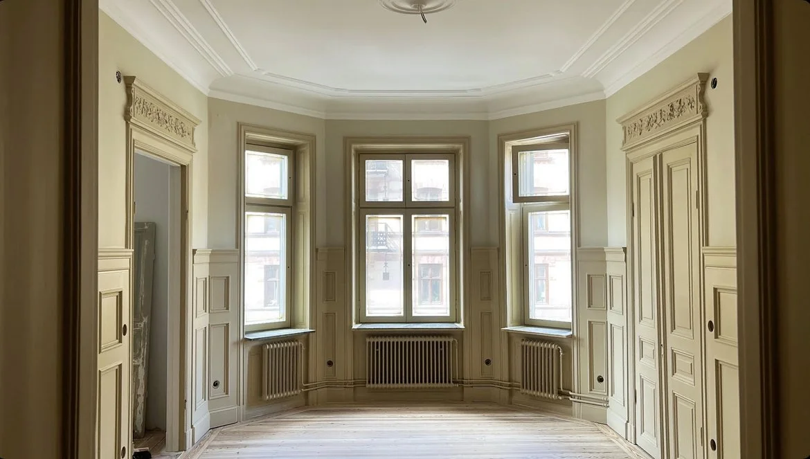 Ett tomt rum med tre stora fönster, detaljerad snickeri och radiatorer under varje fönster.